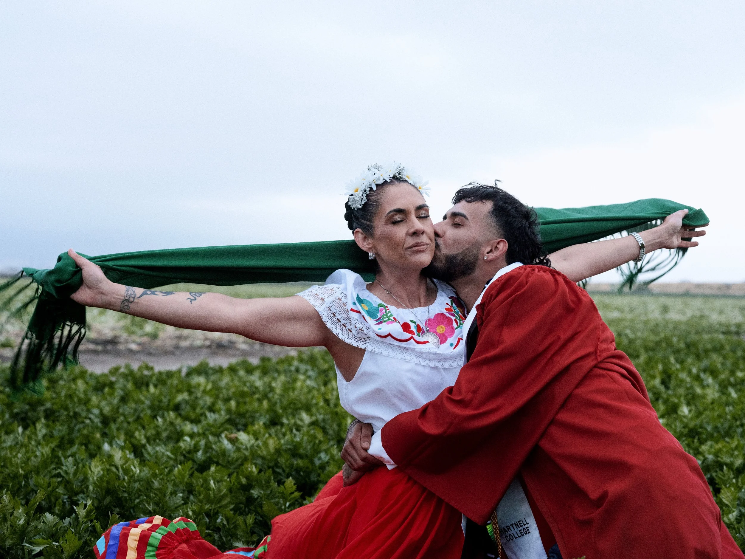 A woman with a floral crown and closed eyes is being kissed on the cheek by a man. They are in an outdoor field with green plants. The woman's arms are outstretched holding a green fabric, and she is wearing a white embroidered blouse and jewelry. Th