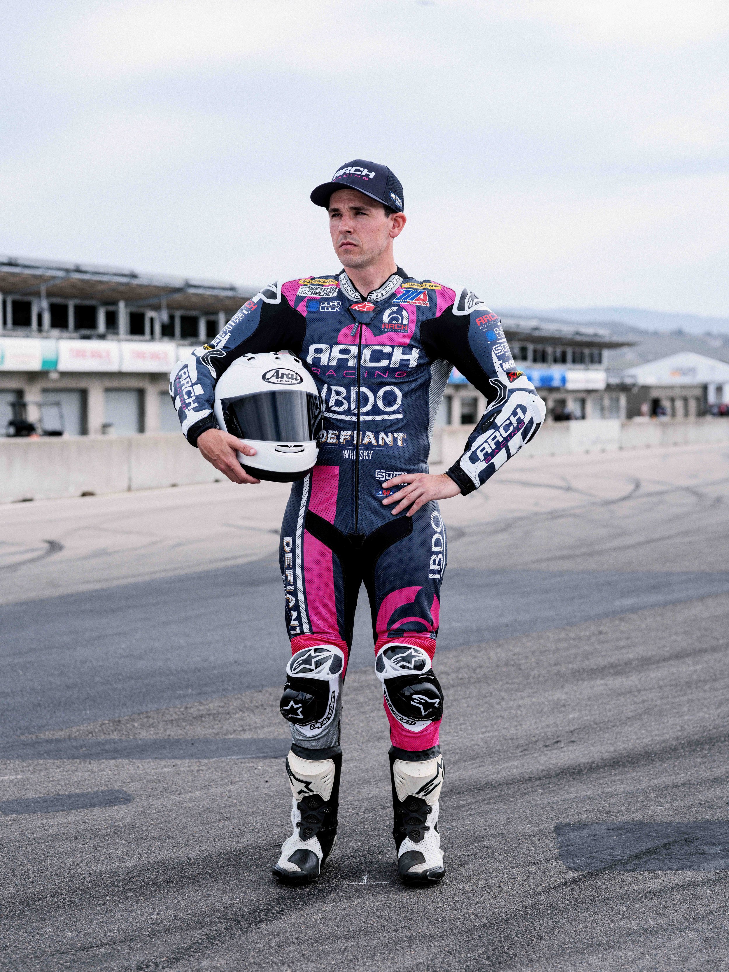 A male motorcycle racer in full racing suit and holding a helmet, standing on a racetrack with grandstands and a cloudy sky in the background.