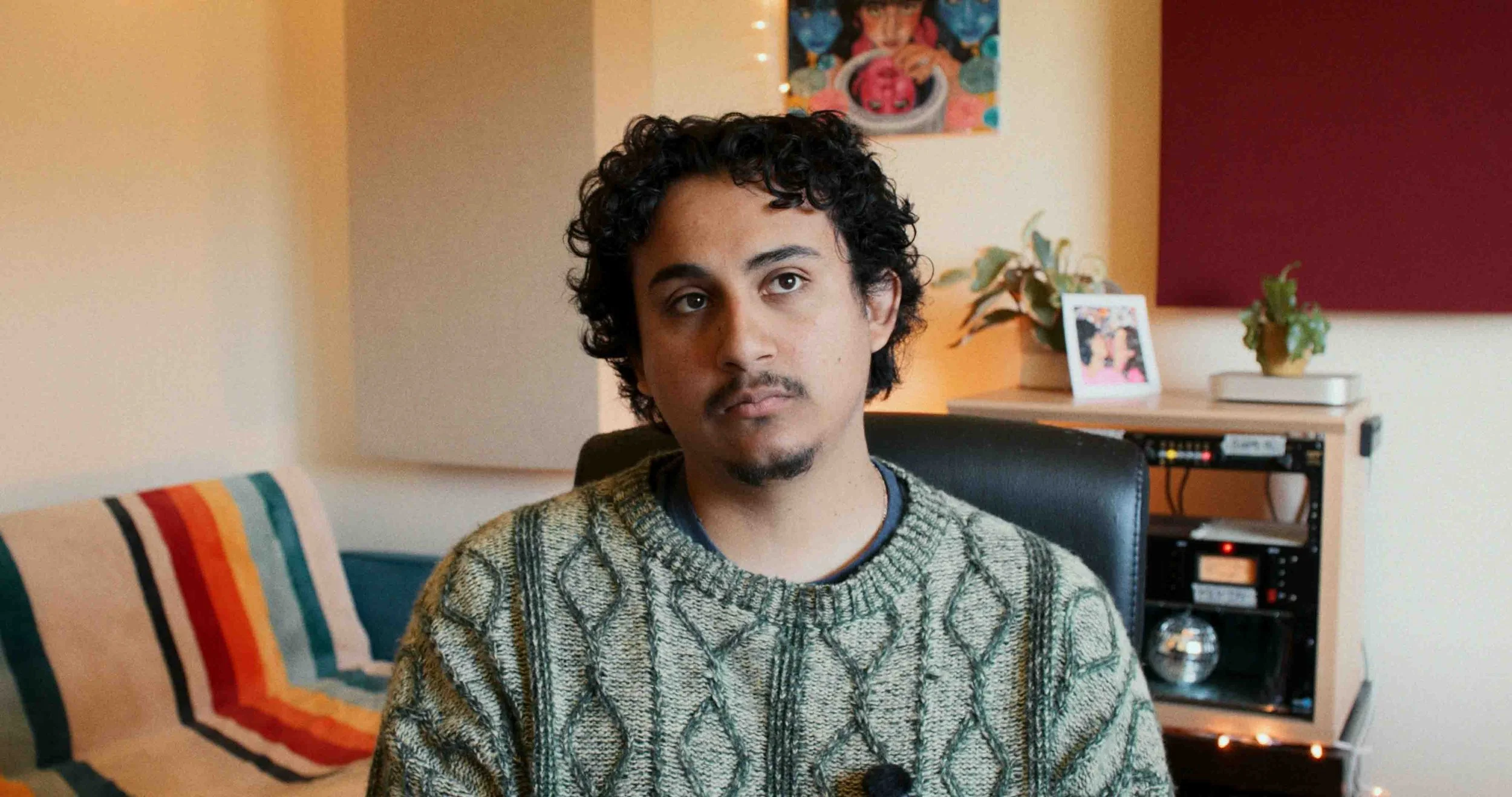 A man with curly dark hair and facial hair, wearing a patterned knit sweater, sitting in an office or living room with a colorful striped pillow, plants, framed pictures, and audio equipment in the background.