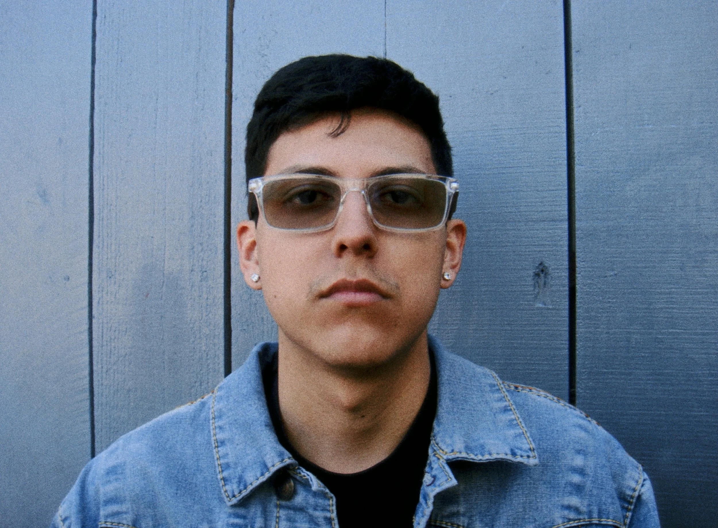 Close-up of a young person with short dark hair, wearing clear glasses, earrings, a denim jacket, and a black shirt, standing in front of a wooden blue wall.
