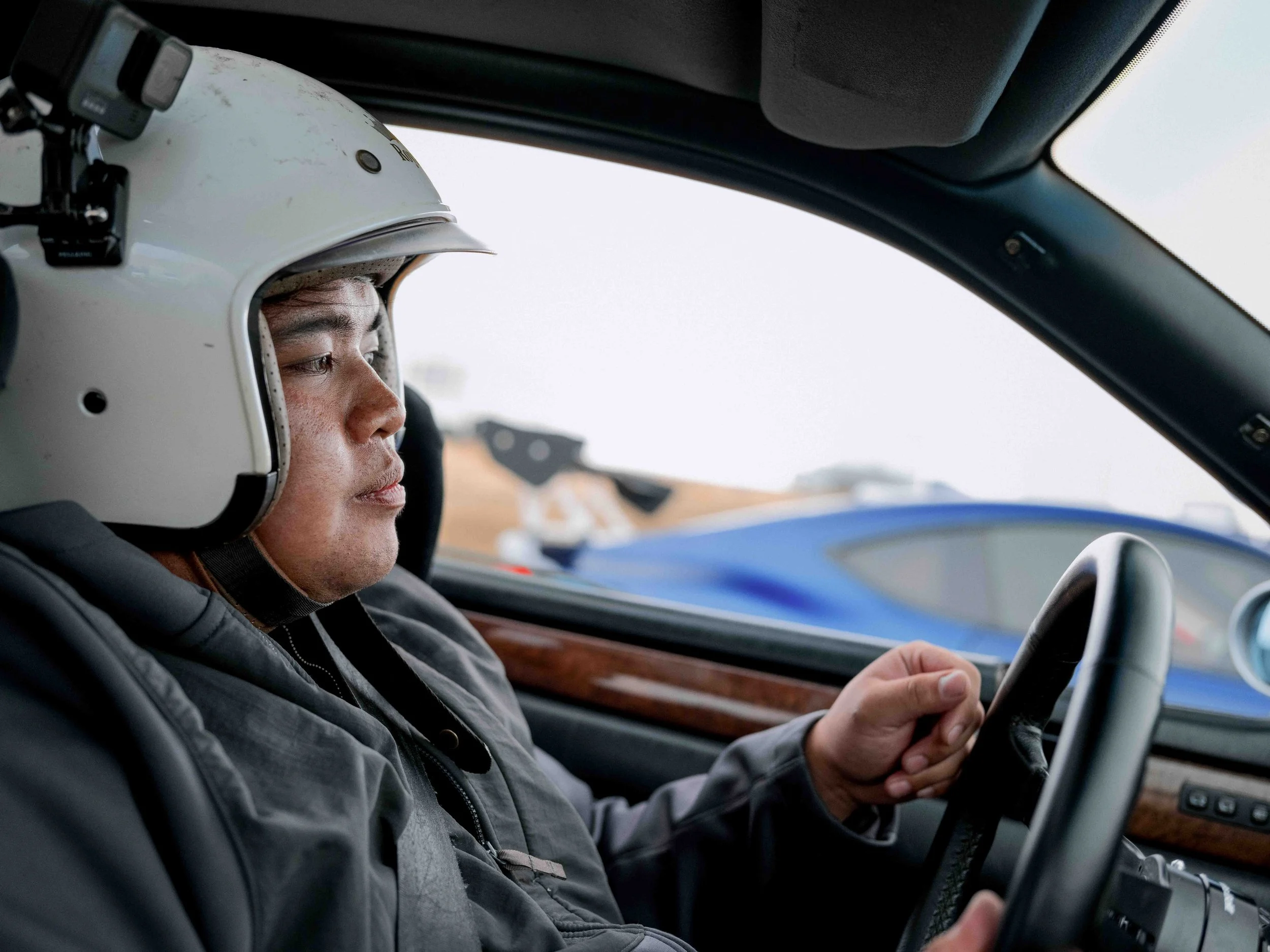 A person wearing a white racing helmet with a black strap, sitting inside a car and holding the steering wheel, with a serious expression.