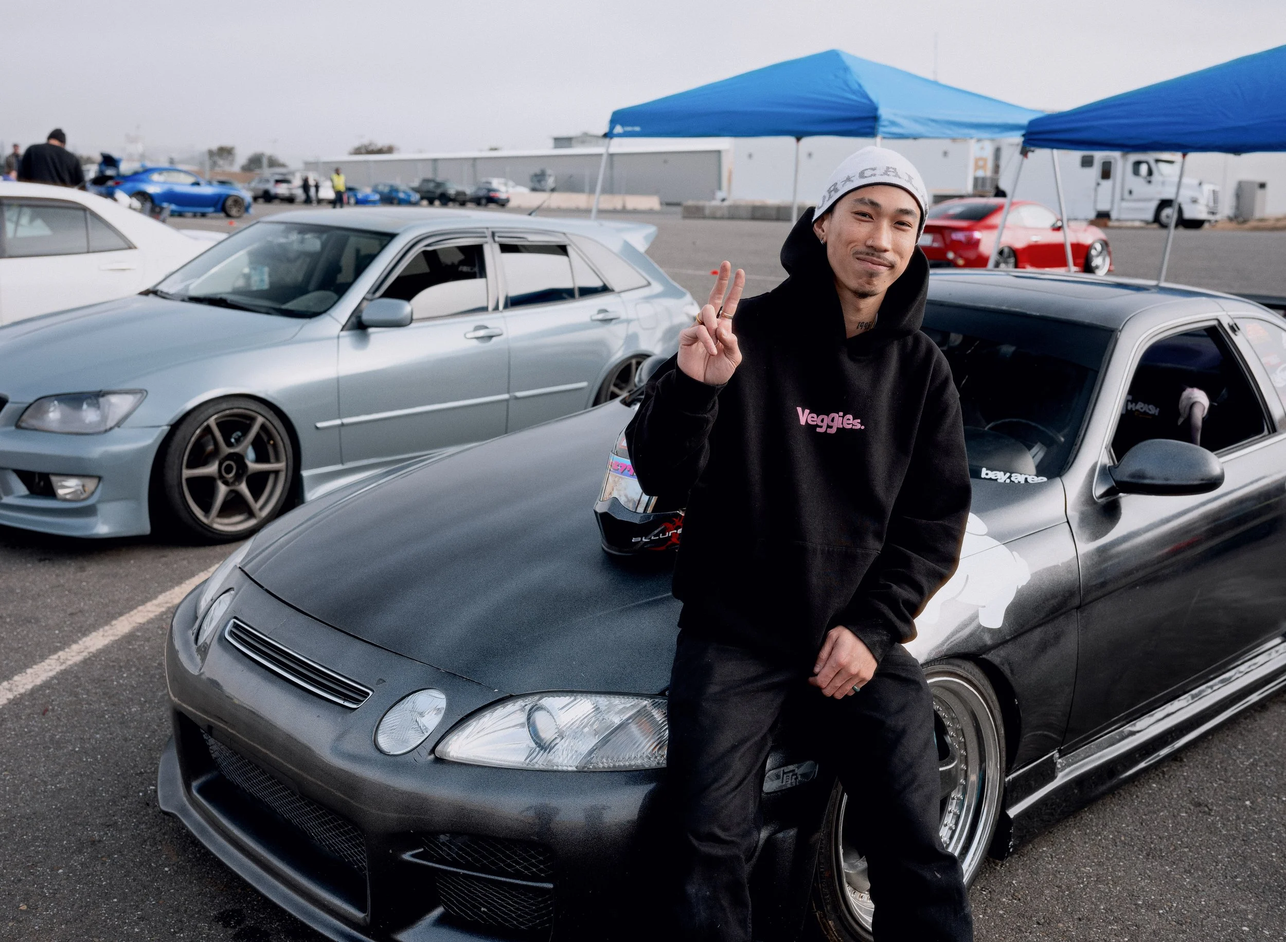 A young man with a peace sign pose standing next to a gray sports car at a car meet, with several other cars and tents in the background.