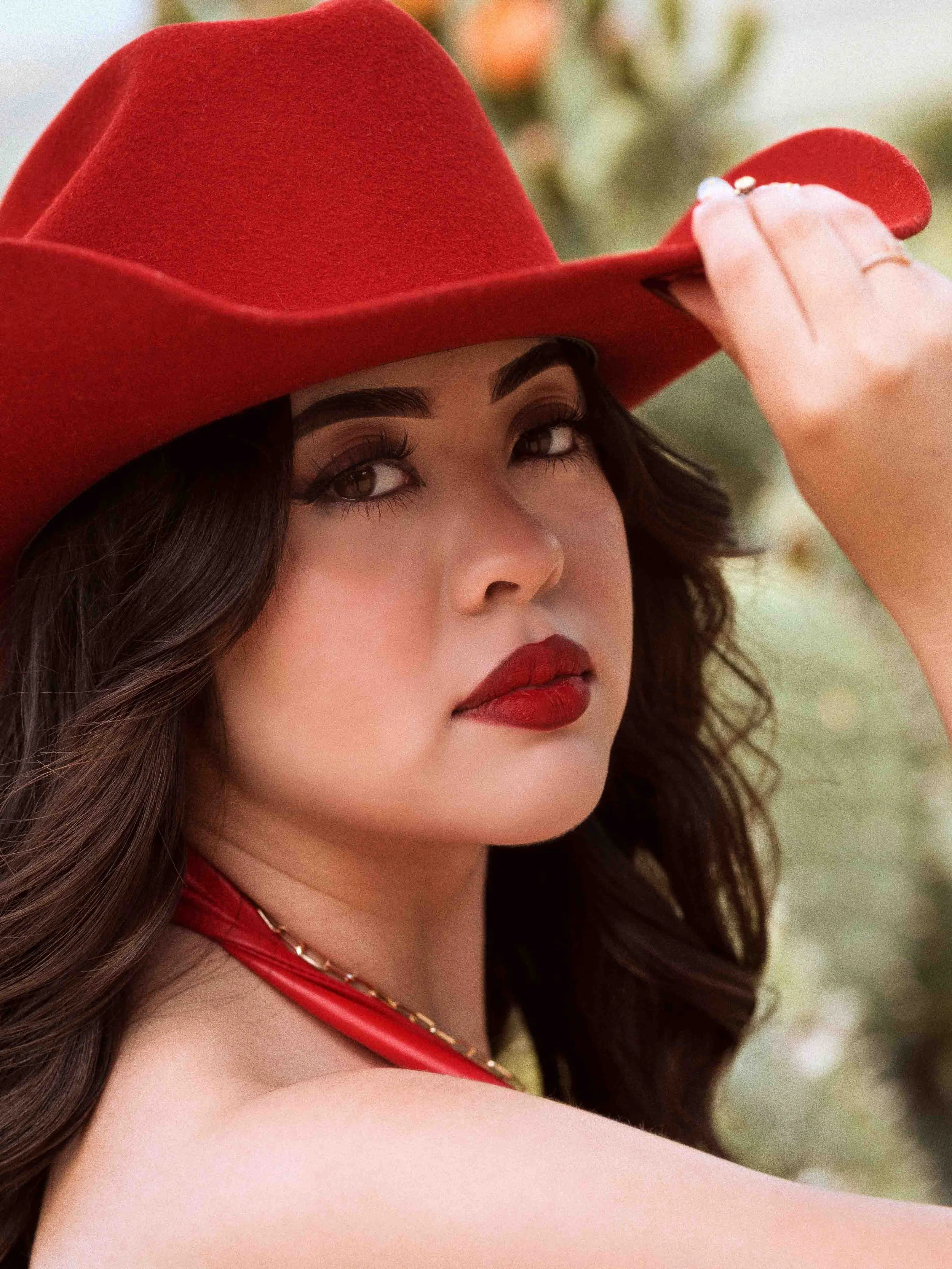 A woman wearing a large red hat, red lipstick, and a red strap necklace, holding the brim of her hat with one hand, with a blurred natural outdoor background.