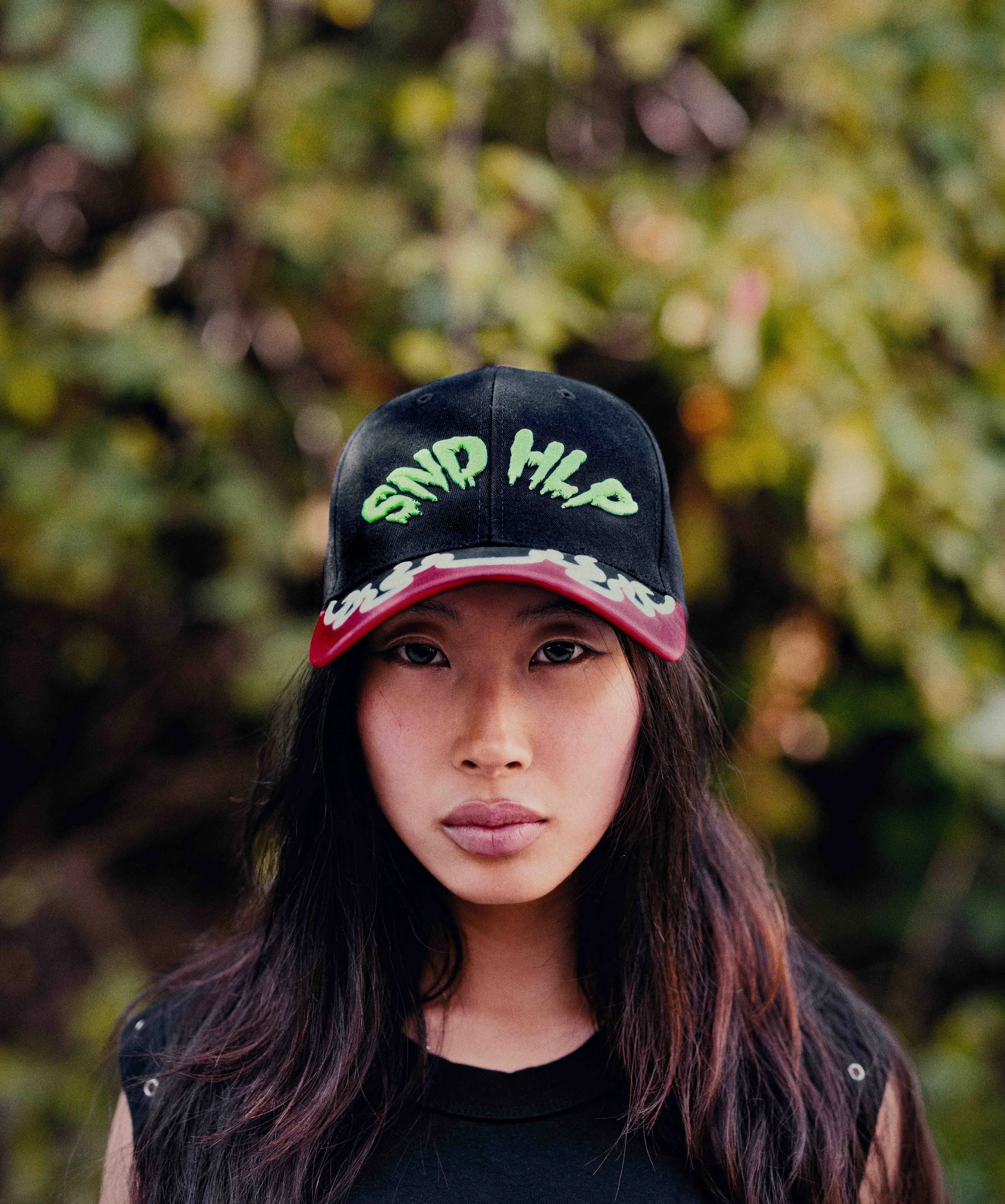 A young woman with long dark hair wearing a black baseball cap with green writing, a red brim, and white trim, and a black shirt, standing outdoors with blurred green foliage in the background.