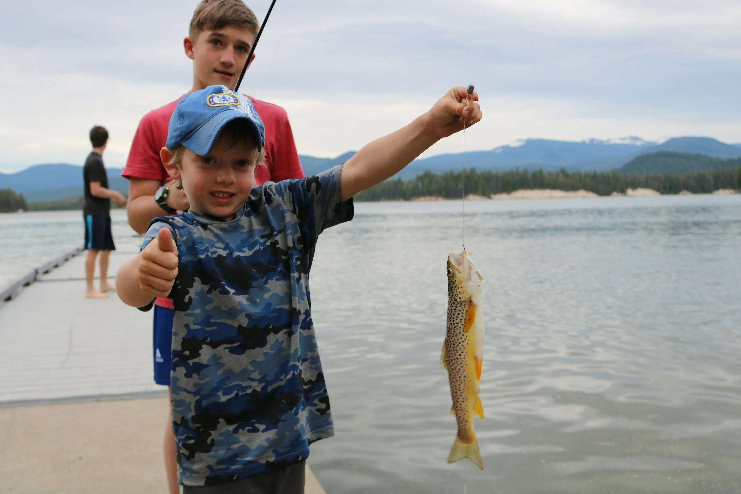 two kids fishing on a lake and smaller kid holding up the fish he caught in one hand and giving a thumbs up with the other hand