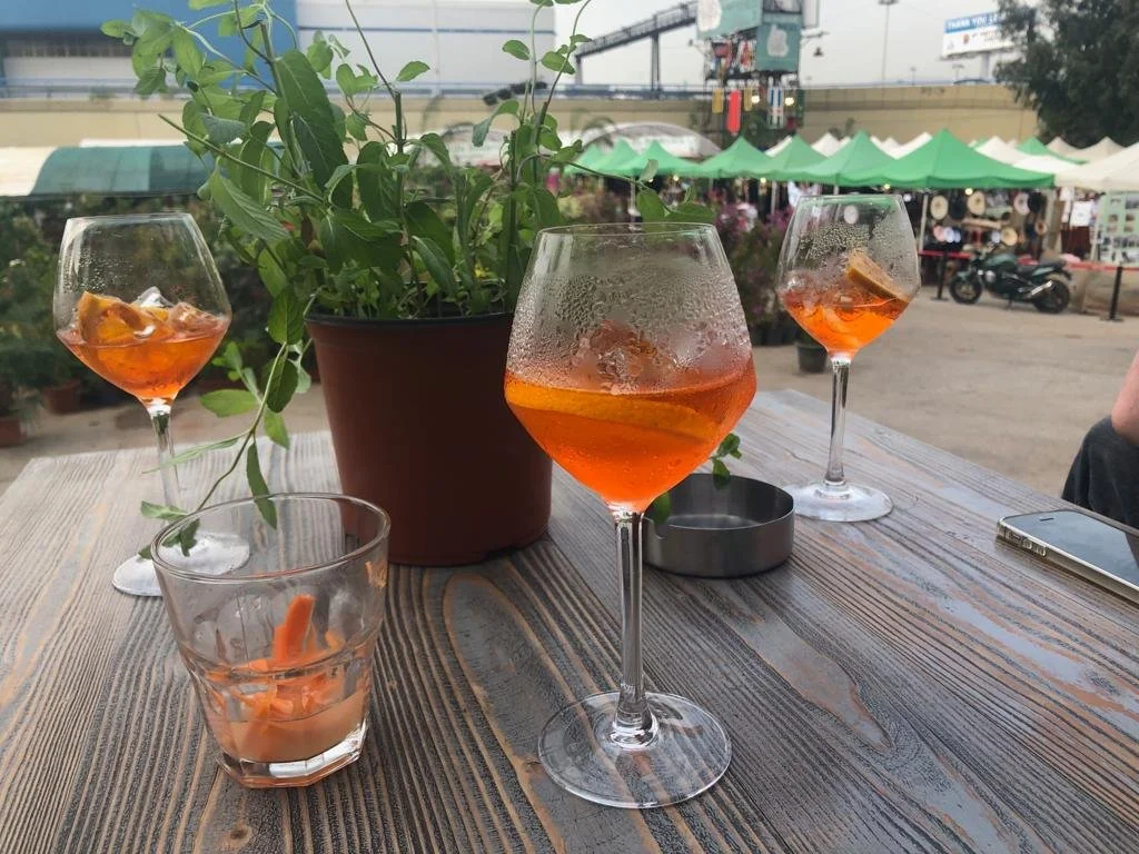 Three glasses of orange beverage with ice, one empty glass with a candle, and a potted plant on a wooden table outdoors at a marketplace.
