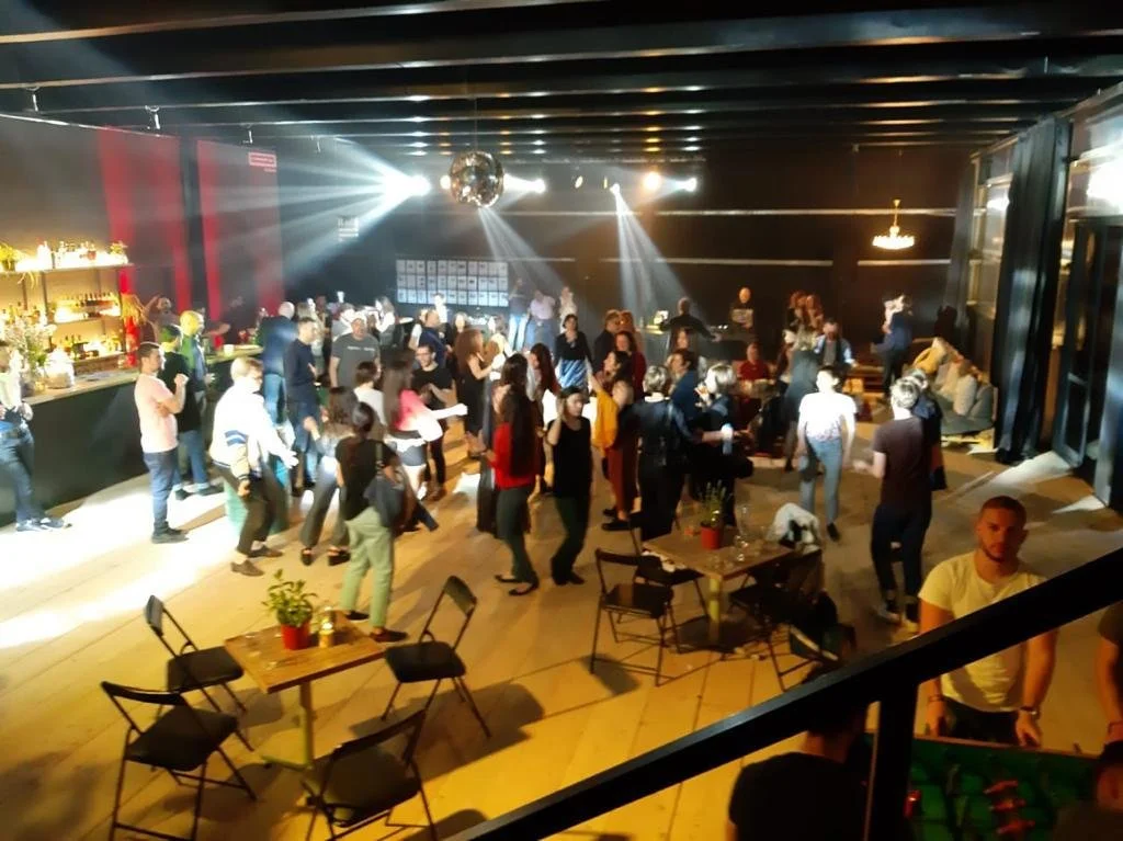 People dancing and socializing in a dimly lit indoor party or club with a dance floor, bar area, and seating.