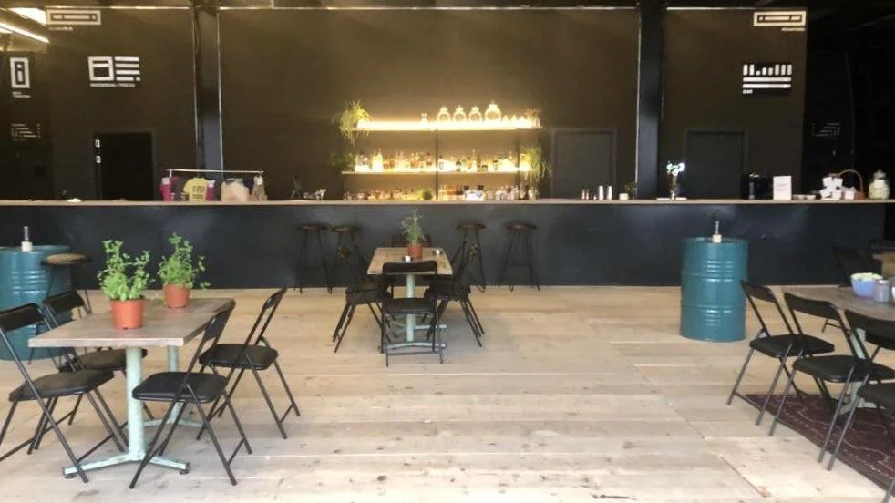Interior of a modern cafe with wooden floors, black walls, tables with chairs, and various plants. Bar area in background with shelves and illuminated decorations.