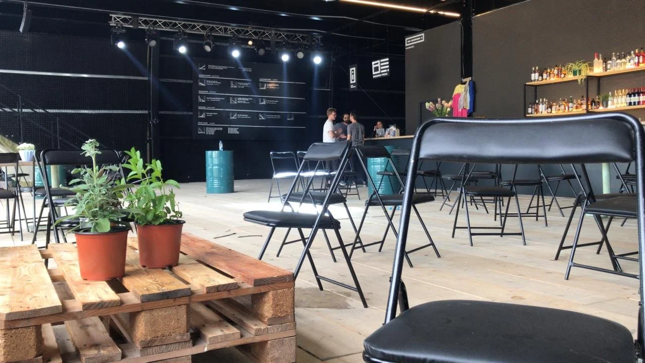 Indoor event space with black walls, stage with lighting, and a bar with bottles. Black chairs are scattered around, some with metal legs. Potted plants on a wooden pallet are in the foreground.