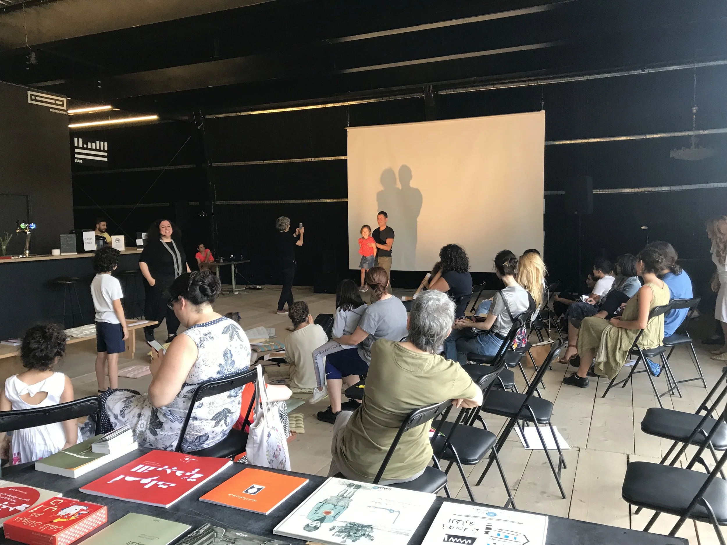 People sitting and standing in a dark room watching a presentation on a large screen. A woman is taking a photo of two children standing in front of the screen, with their shadows cast on it. Books and papers are on a table in the foreground.