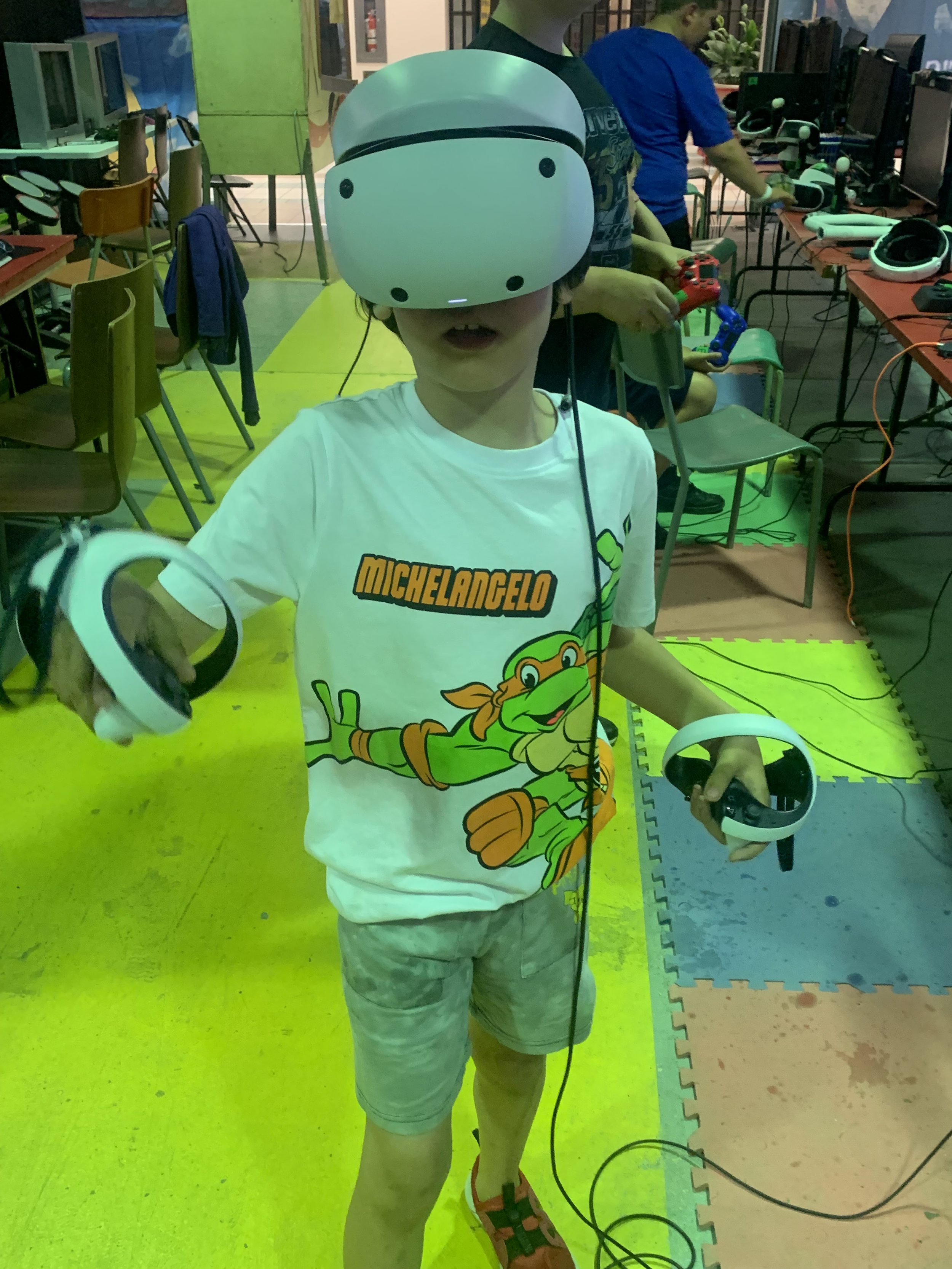 Child wearing a virtual reality headset and holding VR controllers, standing on a colorful foam mat in a gaming or tech center.