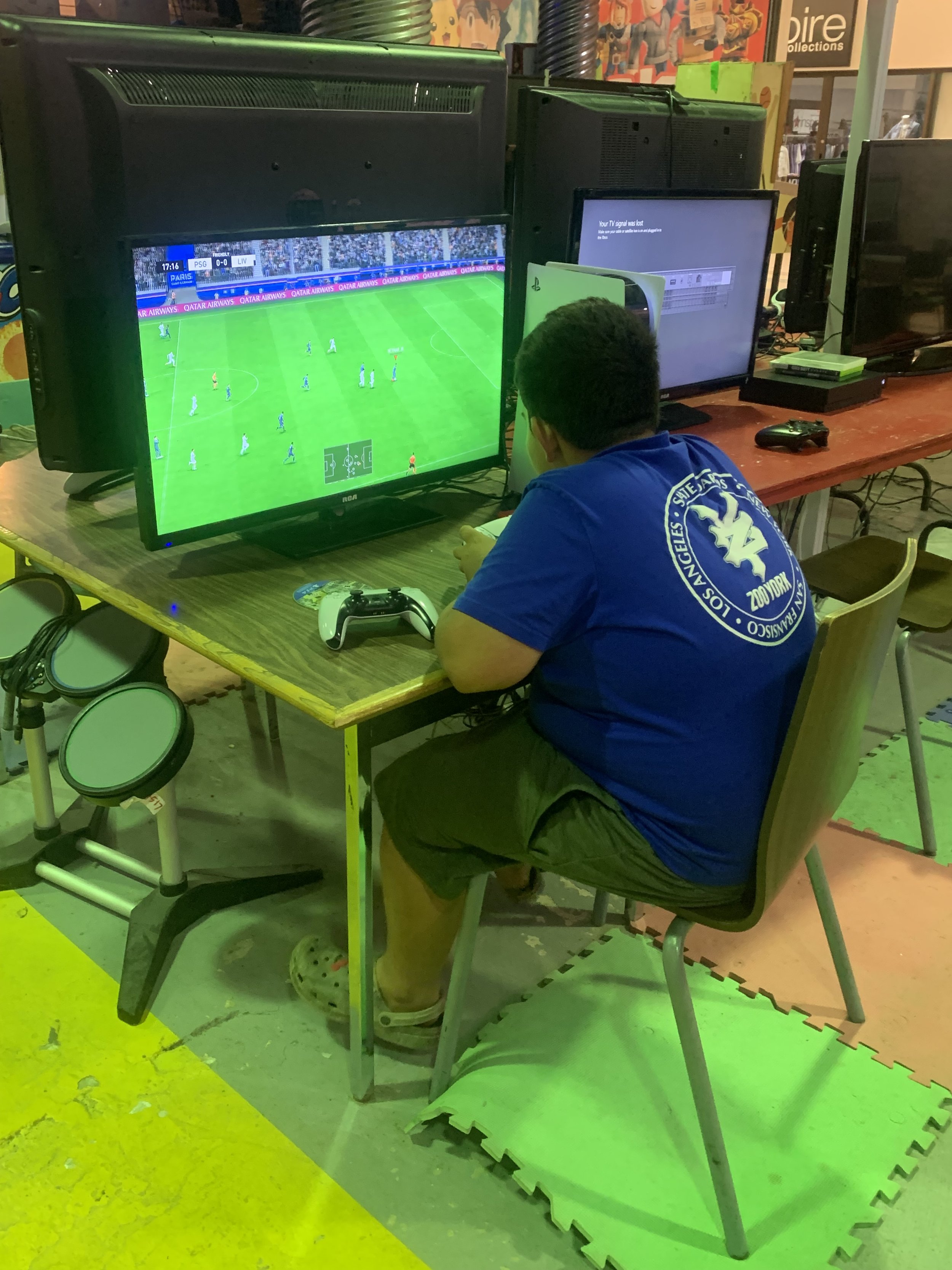 A man playing a soccer video game on a television at an arcade, with multiple monitors and gaming equipment around him.