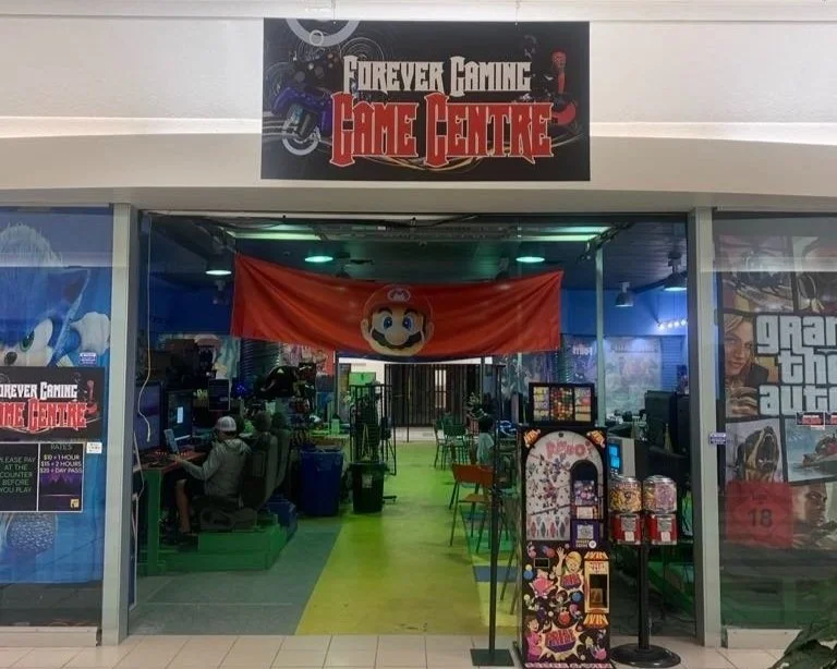 The entrance to a gaming arcade called 'Forever Gaming Game Centre,' decorated with a Mario flag, with people playing video games inside, and arcade machines and posters visible.