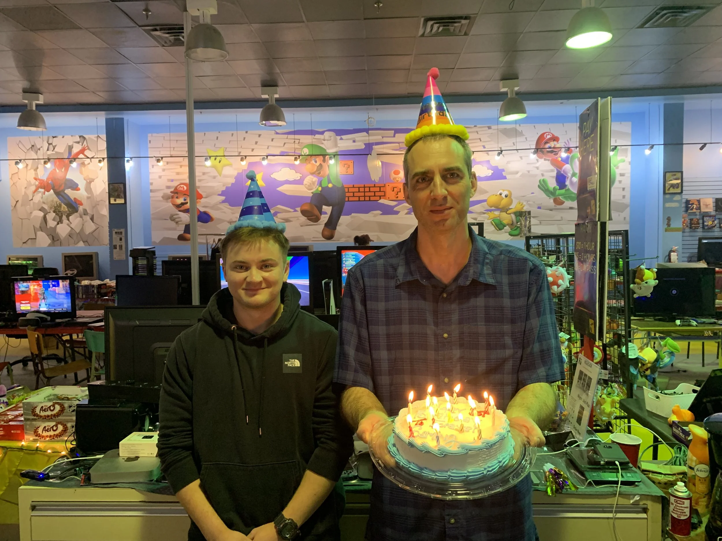 A man and a boy celebrate a birthday in a gaming arcade, wearing party hats, with a cake with lit candles, and gaming arcade decorations in the background.