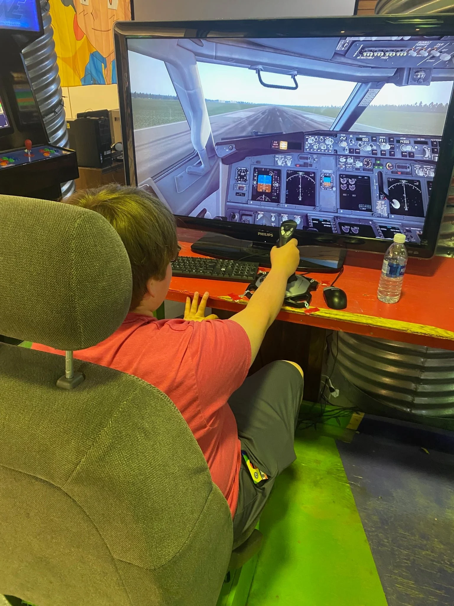 Child sitting in a chair and holding a joystick, playing a flight simulation game on a large screen that displays the cockpit of an airplane on a runway.