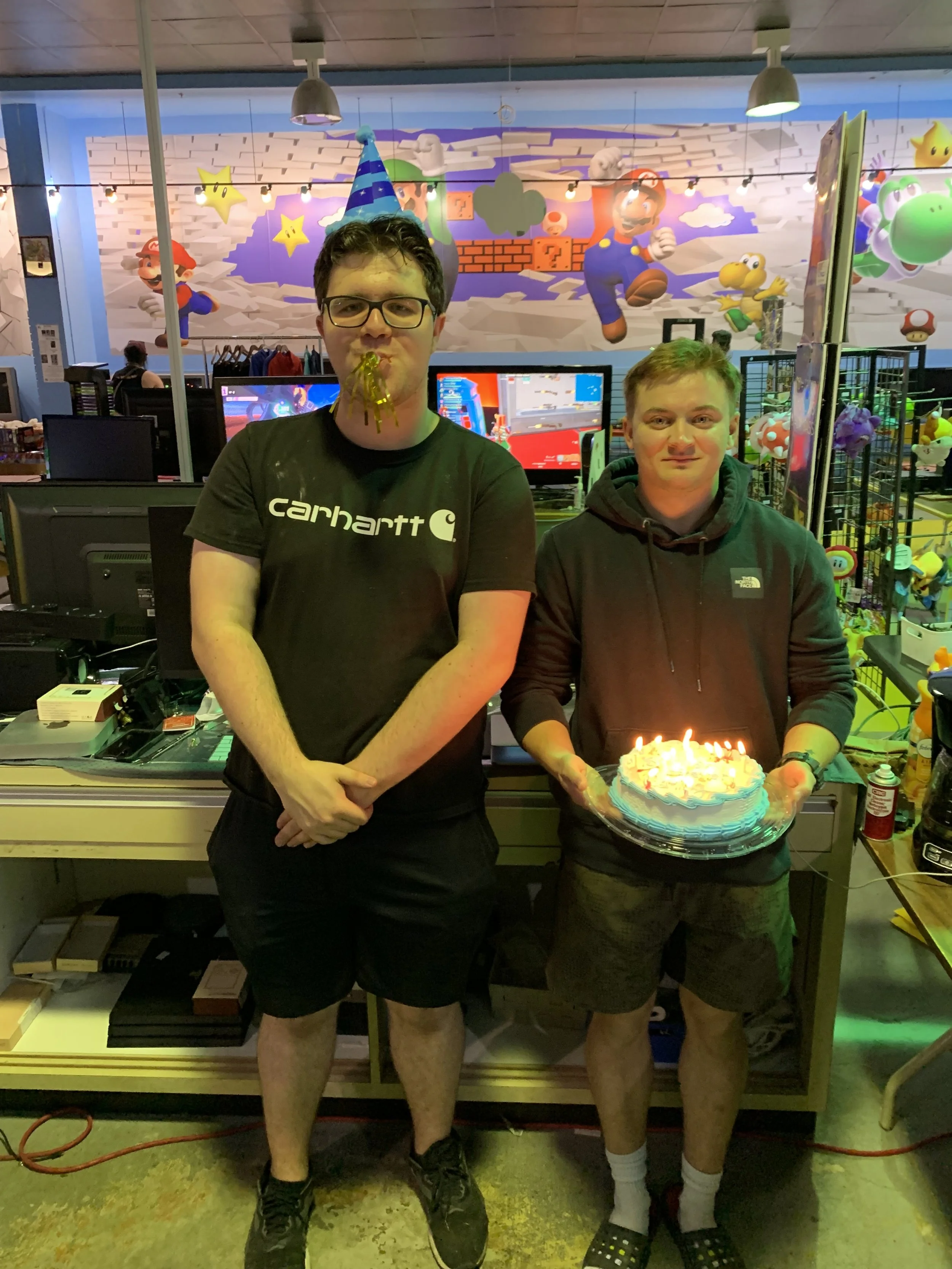 Two young men standing in front of a colorful video game-themed mural, one holding a birthday cake with lit candles, the other wearing a party hat and with spaghetti on his face.
