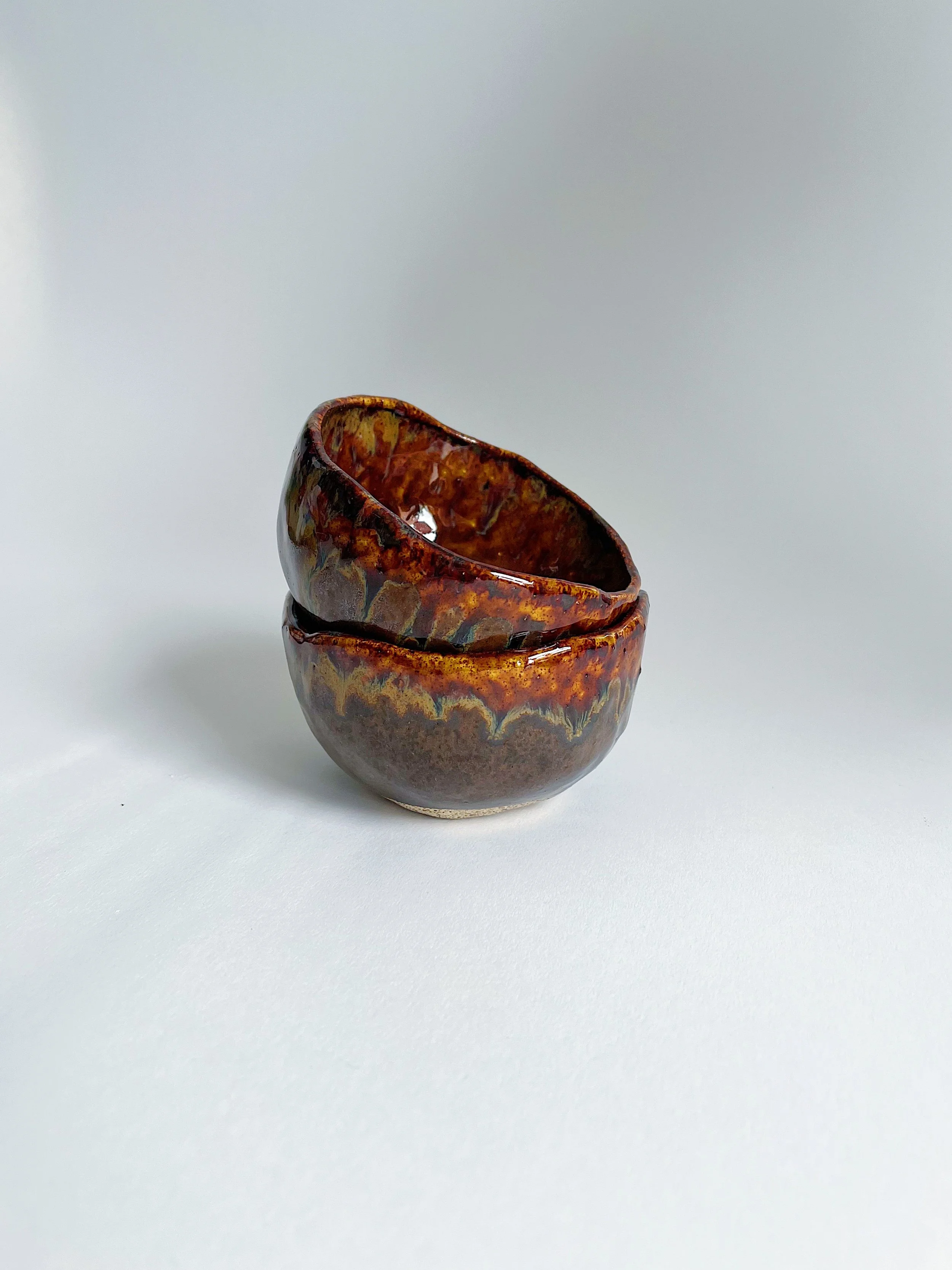 Two small glazed ceramic bowls stacked with a colorful, rustic glaze, placed on a plain white surface against a plain background.