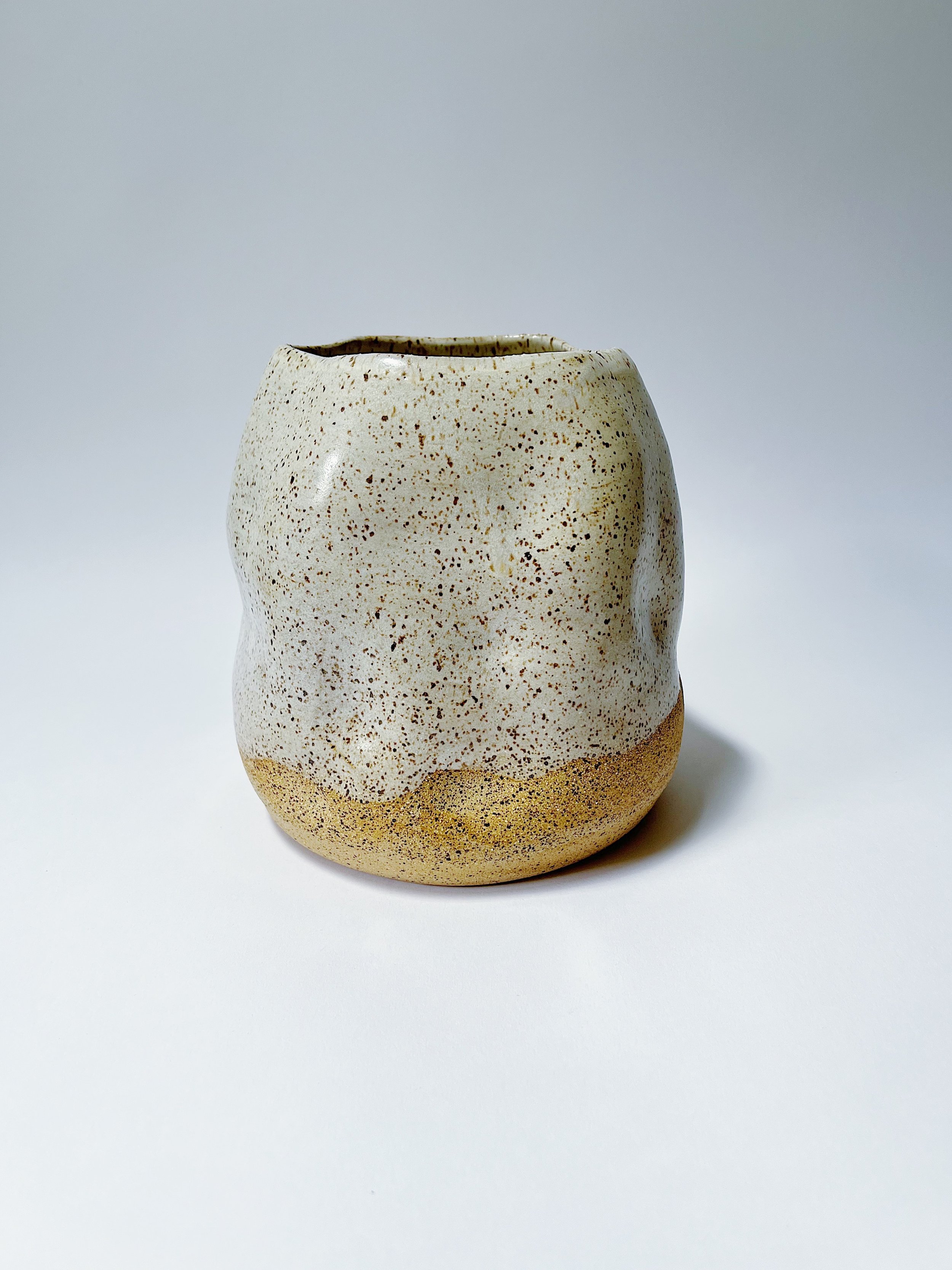 Ceramic vase with a rounded, irregular shape, glazed in a speckled white and tan color, placed on a white surface with a plain white background.