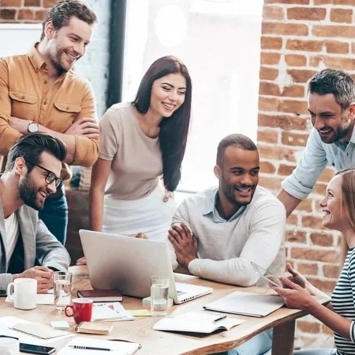 Six people gathered around a table in a modern office, smiling and engaging in conversation. There are laptops, notebooks, and coffee cups on the table. The background features an exposed brick wall and large windows.