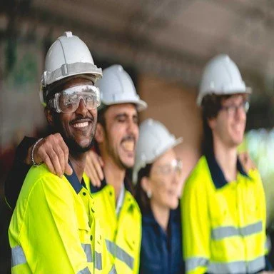 Four construction workers wearing white helmets and high-visibility yellow shirts, smiling together on a construction site.