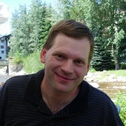 A person smiling outdoors with trees and a river in the background.