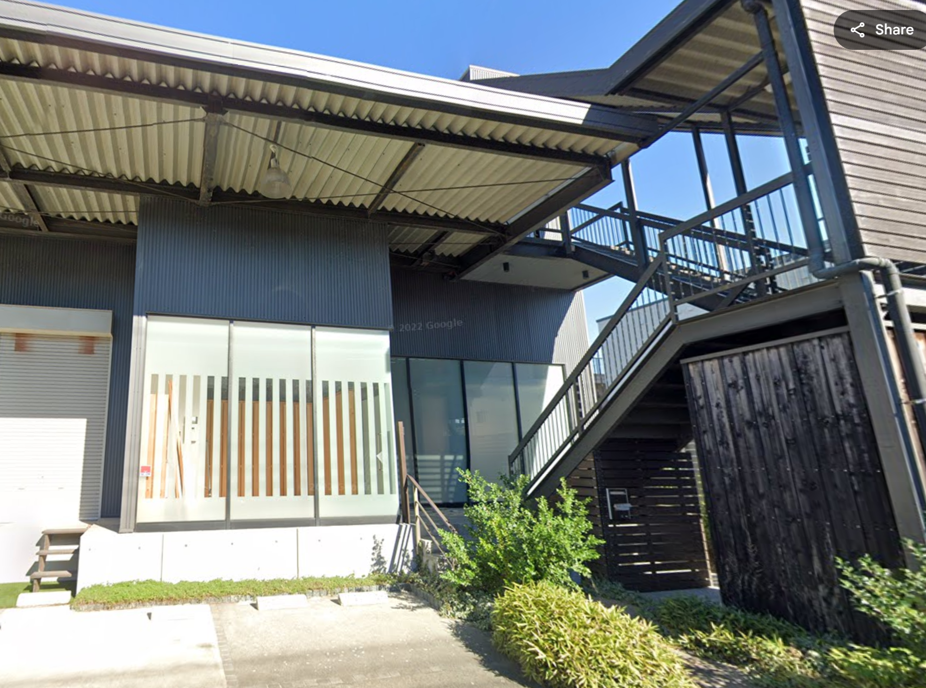 Exterior view of a modern industrial-style building with metal siding, large windows, and an outdoor staircase with black railings, surrounded by greenery and a parking area.