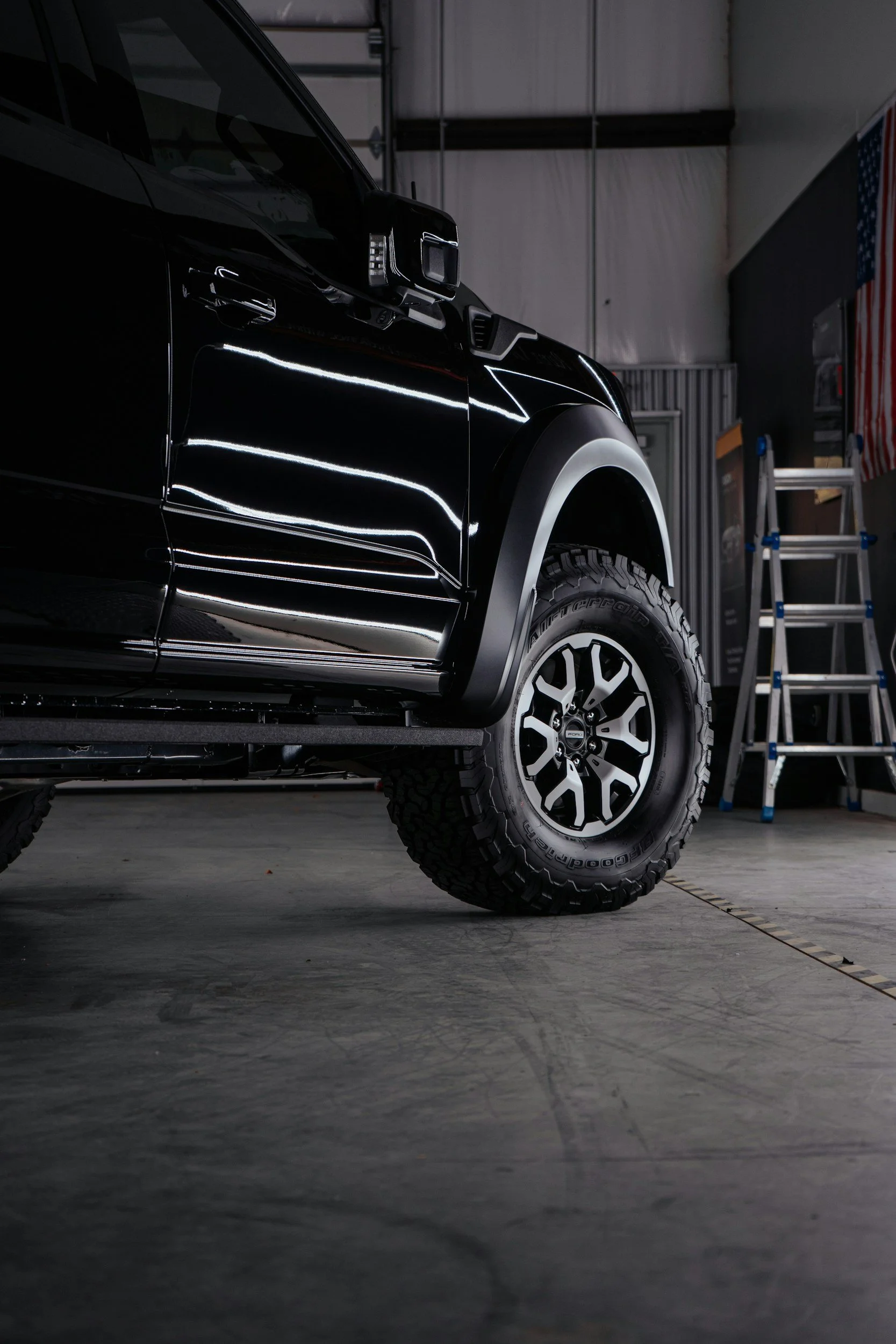 Close-up of the front side of a black pickup truck with off-road tires and a chrome wheel, parked inside a garage with ladders and an American flag in the background.