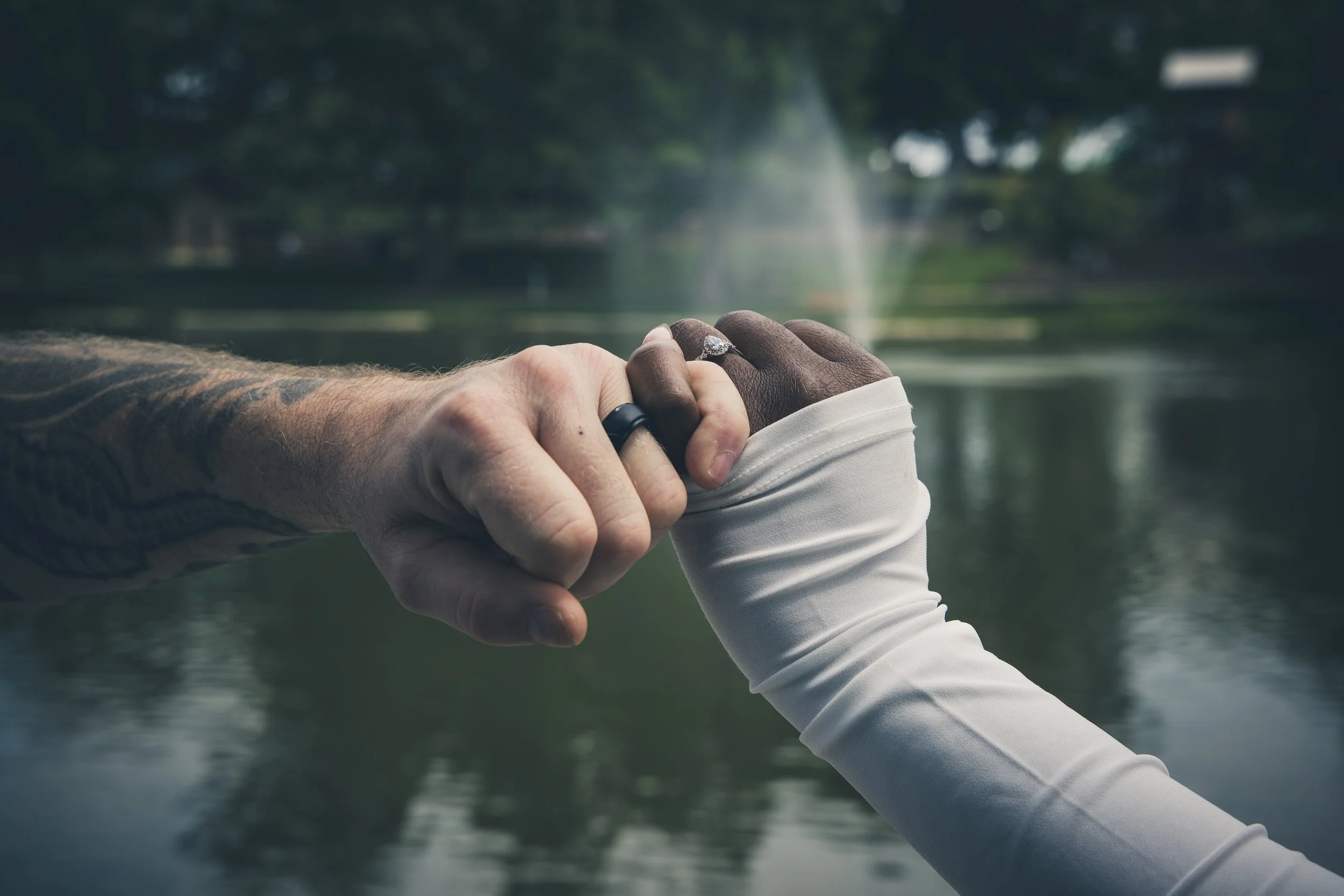 couple hands with wedding bands pinky promising with pond and fountain in the background.
