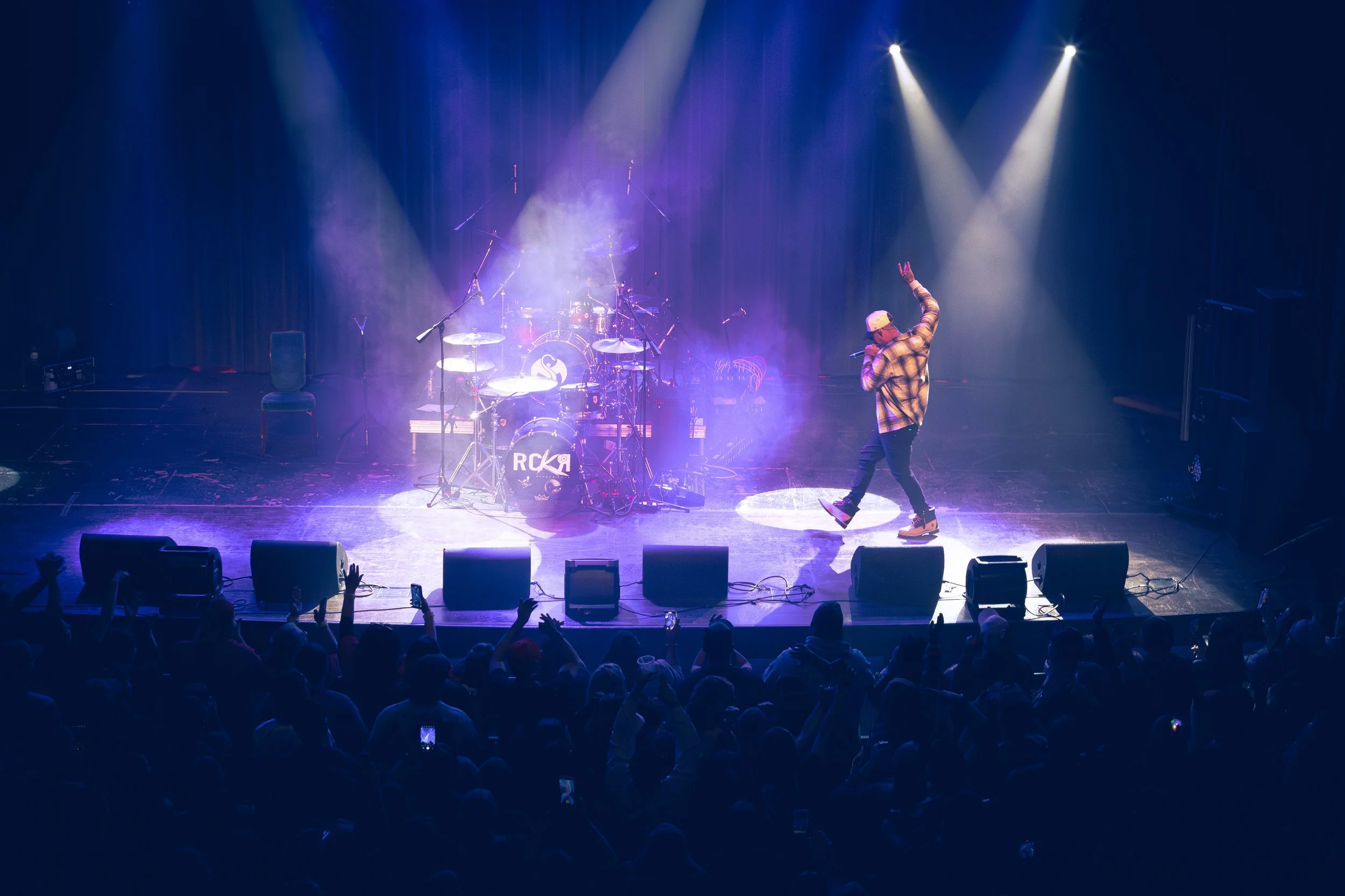 A performer on stage with a microphone, wearing a baseball cap and plaid shirt, dancing near a drum set in front of an audience in a dark concert hall with purple and white stage lighting.