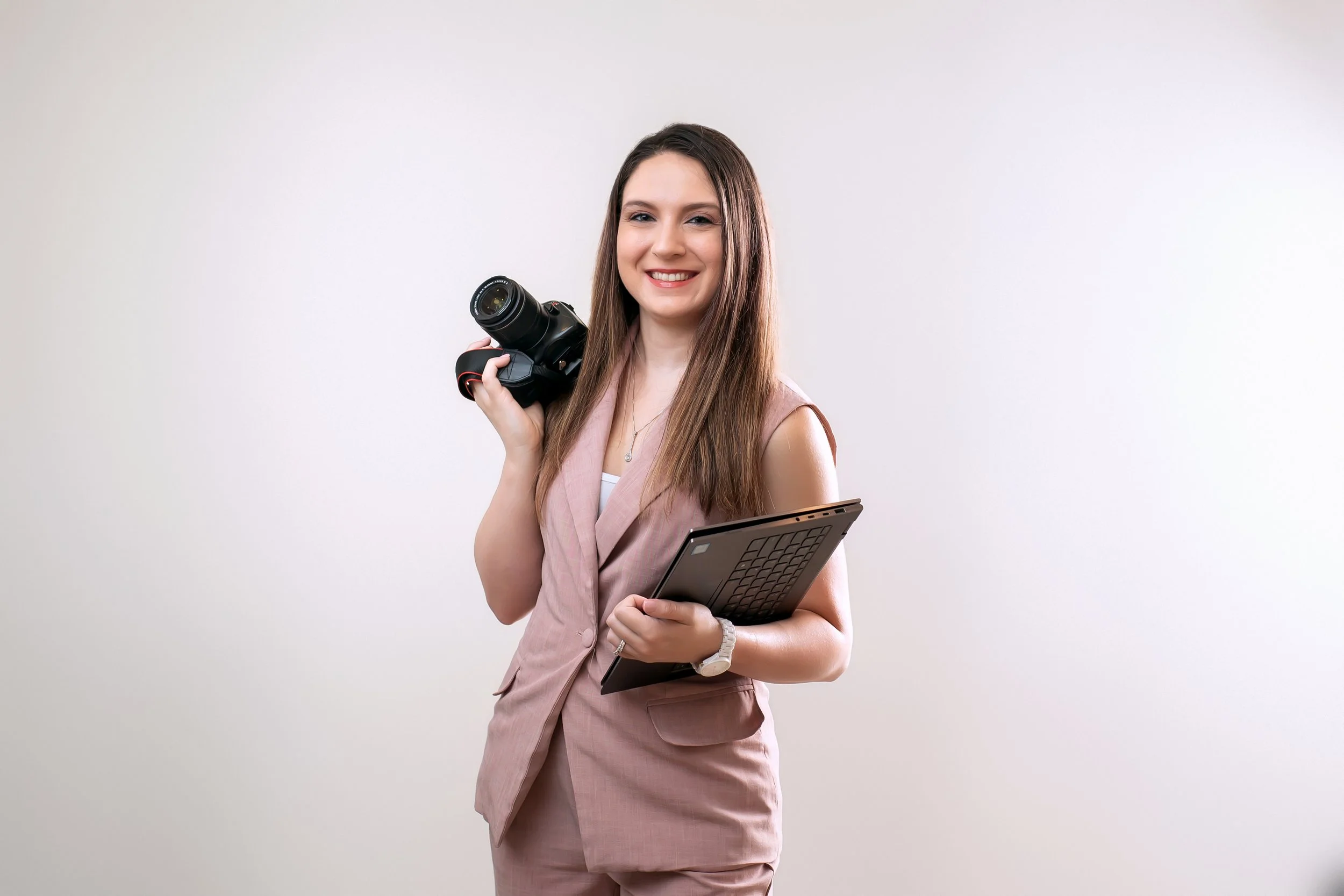 A young woman smiling, holding a camera in one hand and a laptop in the other, standing against a plain white background.