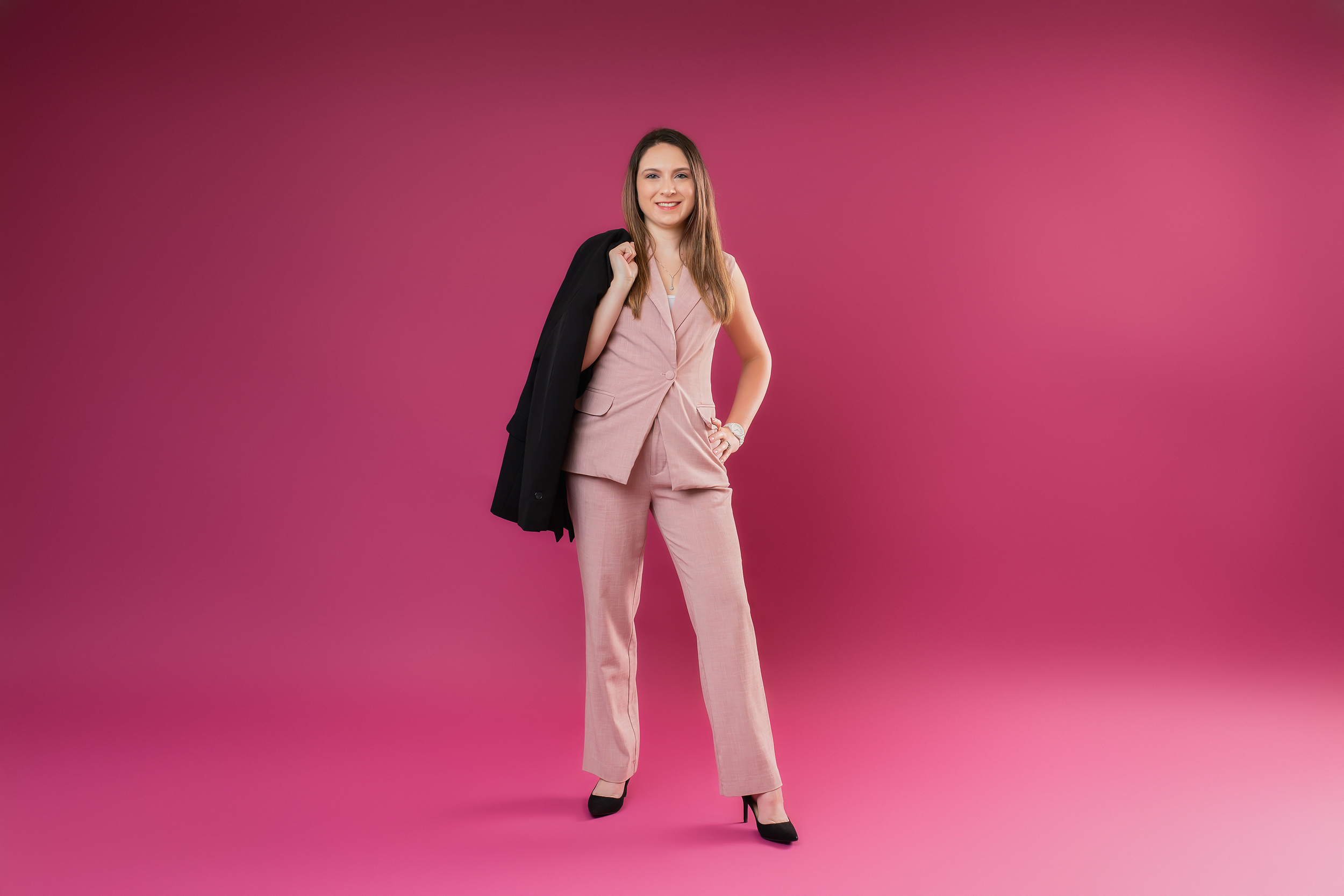 A woman in a pink pantsuit holding a black blazer over her shoulder, standing against a pink background.
