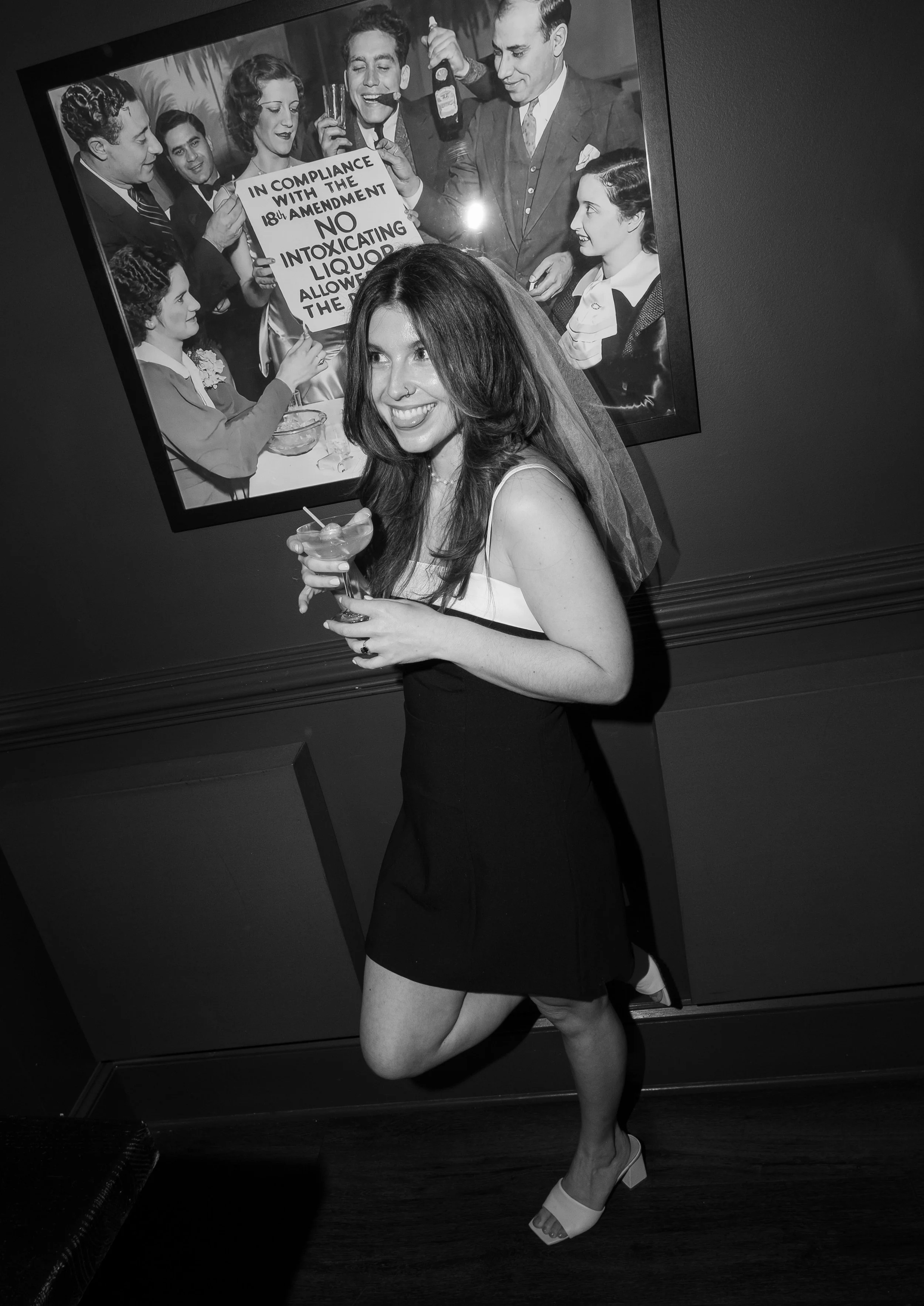 A young woman with long hair and a dress, holding a cocktail, smiling and posing in front of a black-and-white framed photo of a vintage gathering with people holding signs.
