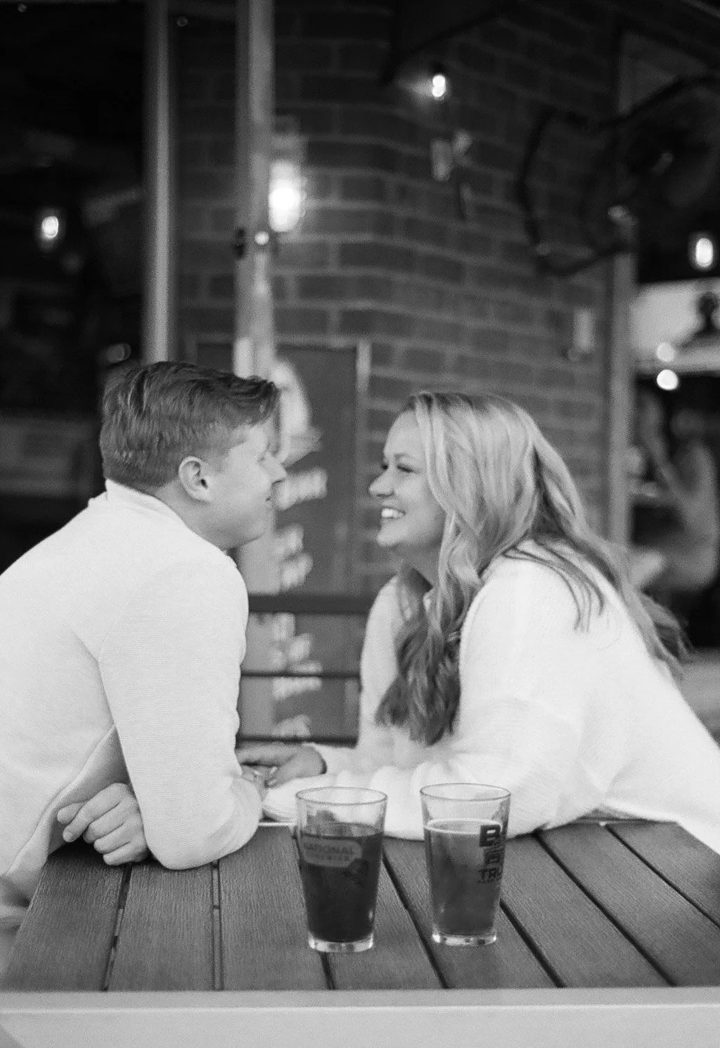 A man and woman are sitting at a table in a restaurant, leaning towards each other and smiling. Two glasses with drinks are on the table.