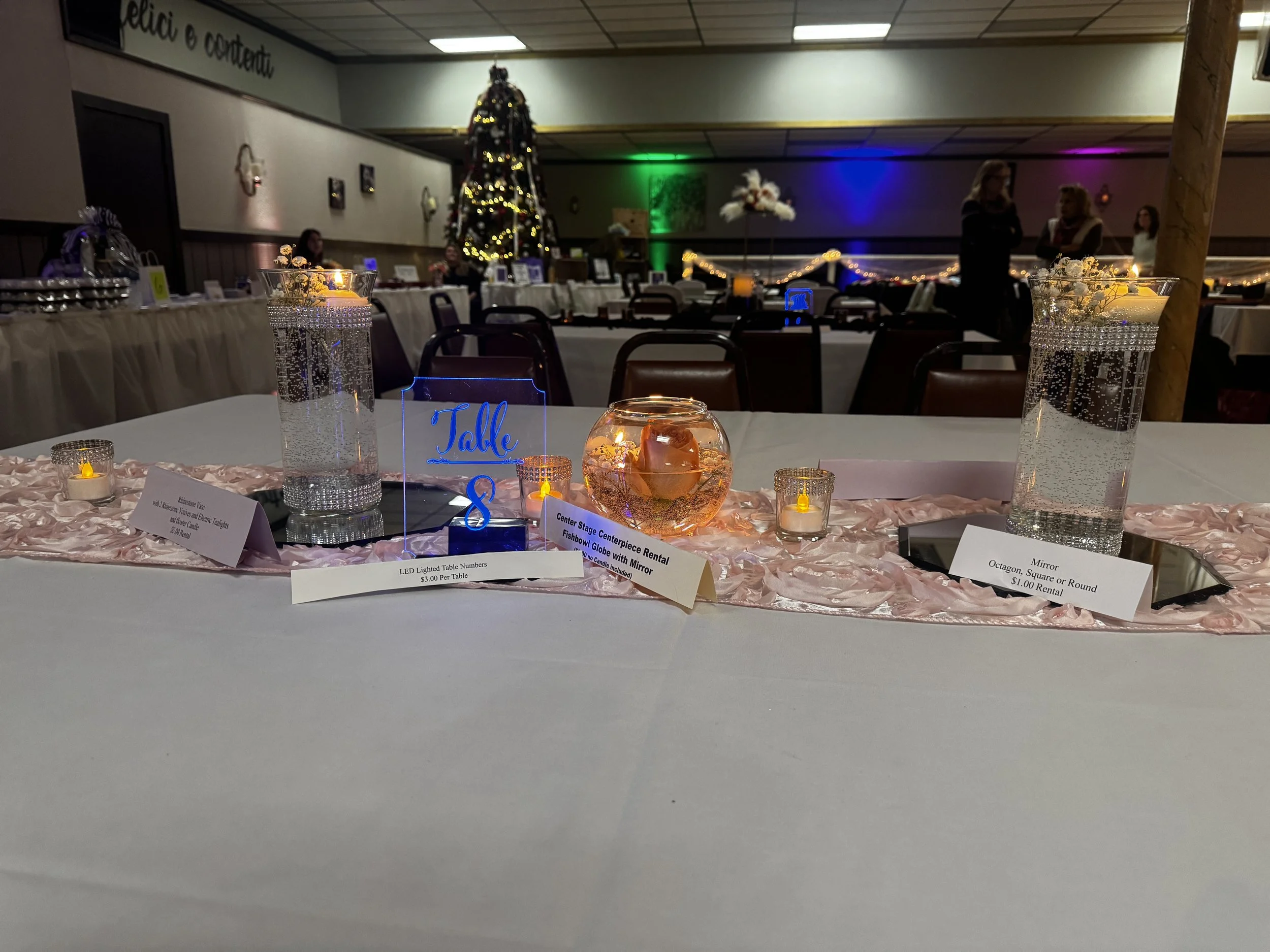 A table decorated at Center Stage with a baby pink runner, two octagon mirrors with vases and floater candles, a globe with a floater candle and a floating rose, votive candles with electric candles, and a lighted table number.
