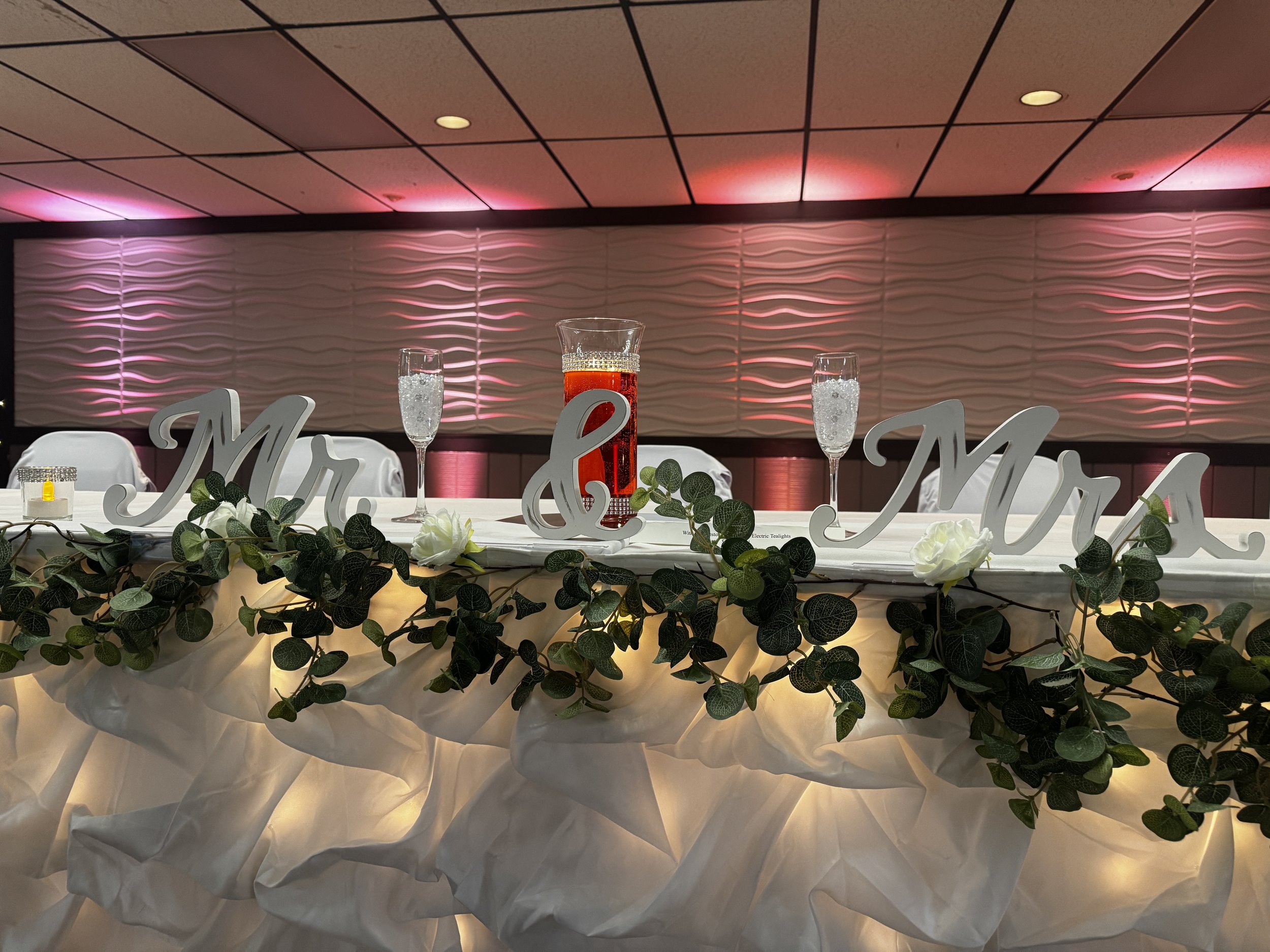 A head table set up for a bridal party. The table is decorated with a Mr. & Mrs. sign, as well as champagne glasses and a vase with colored water and a floater candle. 