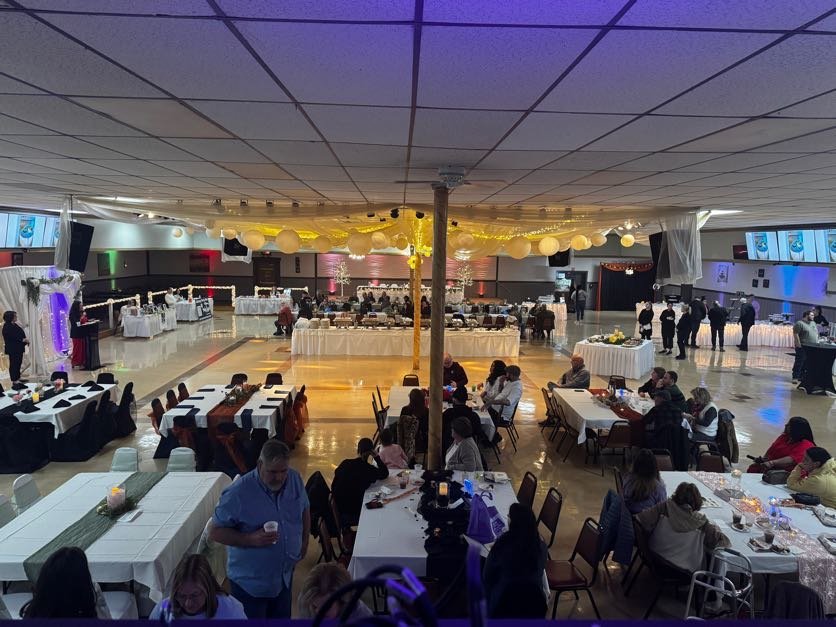 A wide shot photograph of the entire hall of Center Stage, showing multiple tables with various decorations, people sitting at the different tables, and a woman speaking at a podium. Workers stand in the corner, wearing all black, near the buffet. 