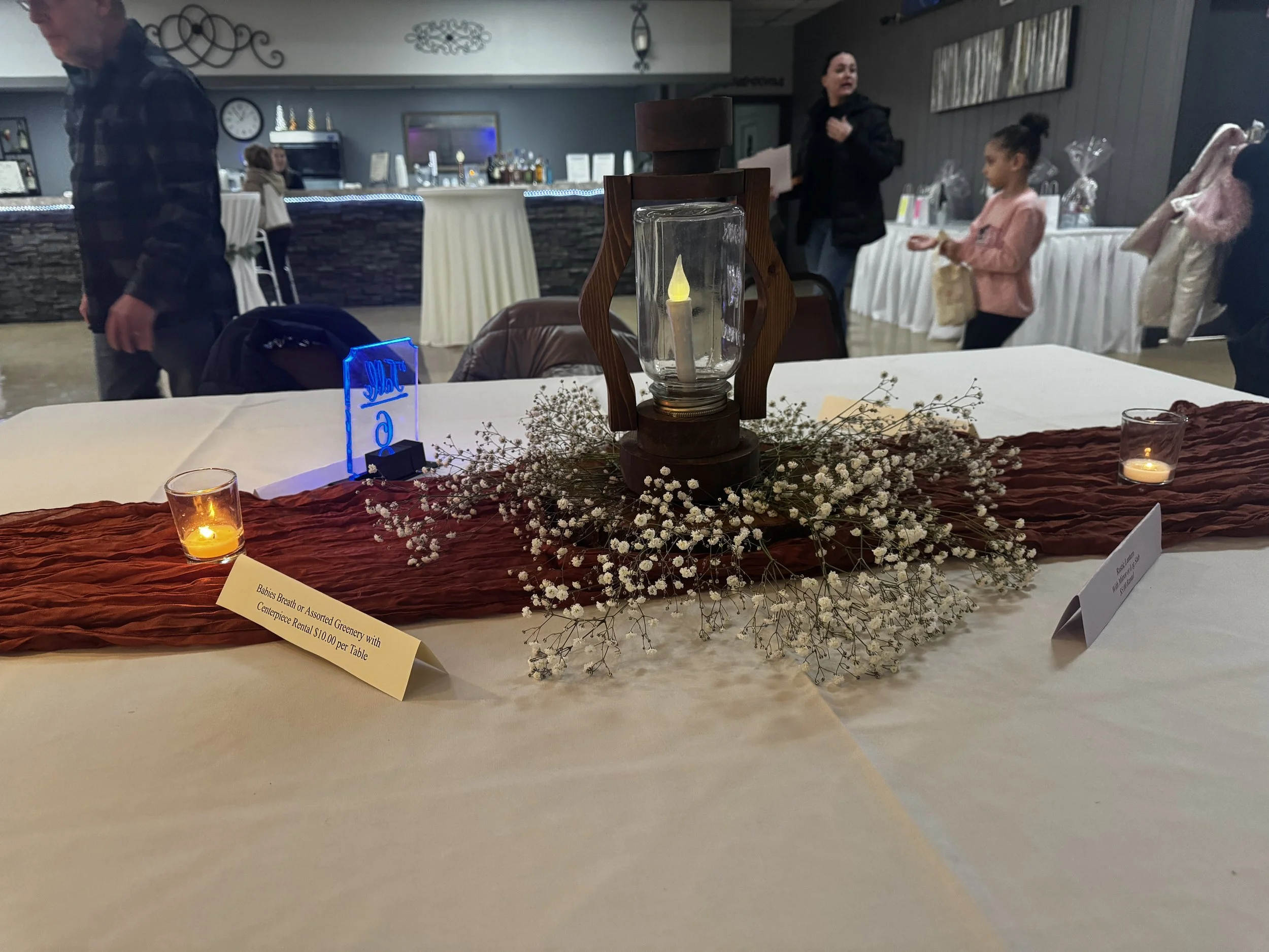 A centerpiece decoration with a burgundy table runner, votive candles, a lighted table number, and a lantern with baby's breath florals underneath. 