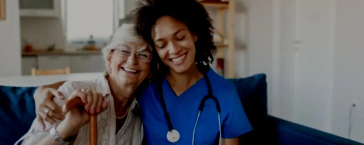 A young female healthcare worker in blue scrubs and a stethoscope hugging an elderly woman with white hair and glasses, both smiling.