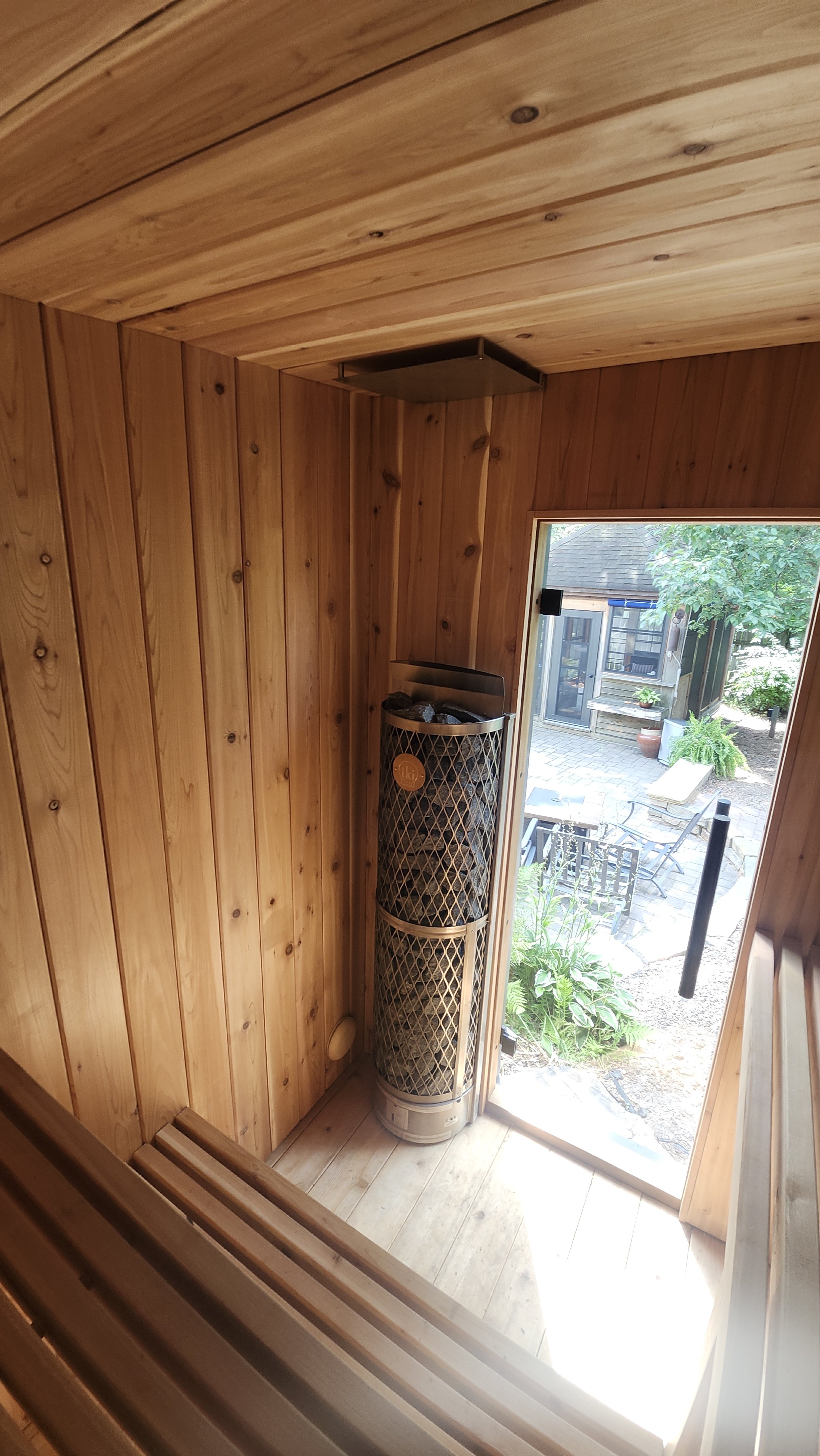 Wood-paneled sauna interior with a heater and an open door leading to a garden patio.