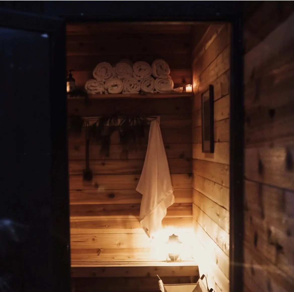 A cozy sauna with wooden walls, a shelf with rolled towels, a hanging towel, candles, and a lantern providing warm lighting.