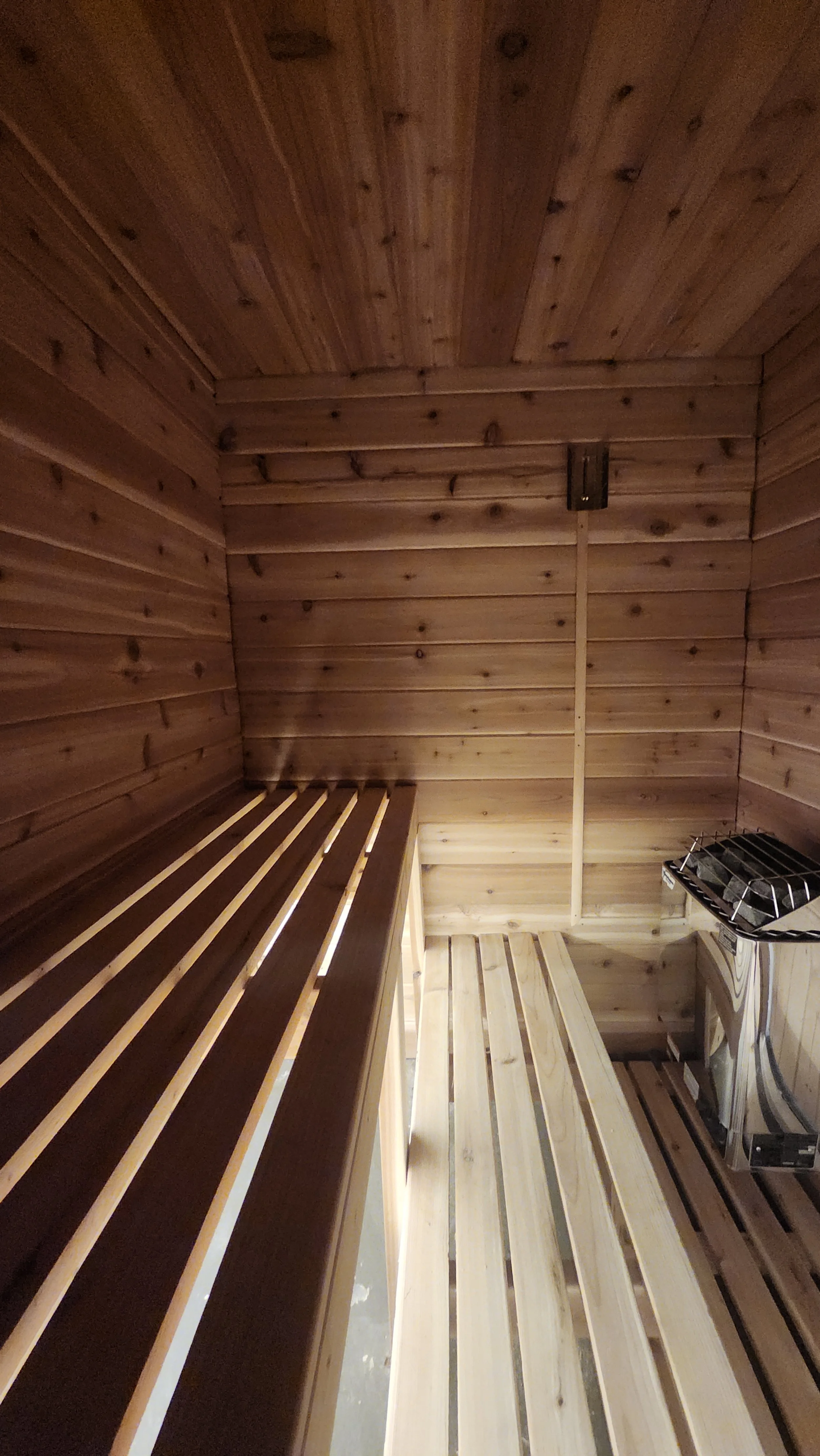 Interior view of a wooden sauna with two benches, a small heater on the right, and wood-paneled walls and ceiling.