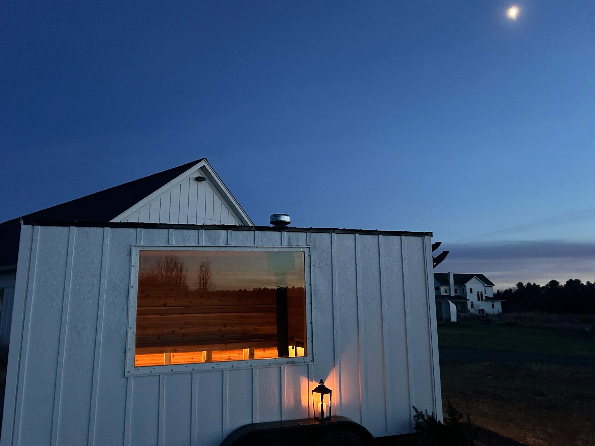 A small white building with a window reflecting trees during twilight, with a lantern light on and a larger house in the background, under a darkening sky with the moon visible.