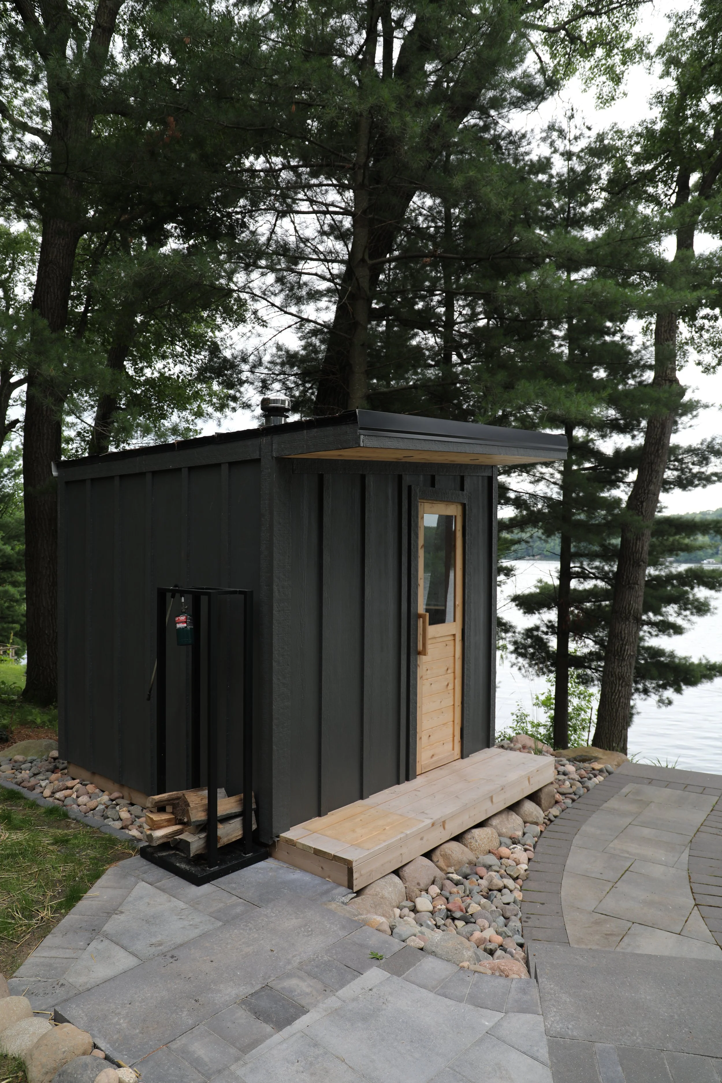 Small black wooden shed with a natural wood door, situated on a stone and paved path near a wooded lakeside.