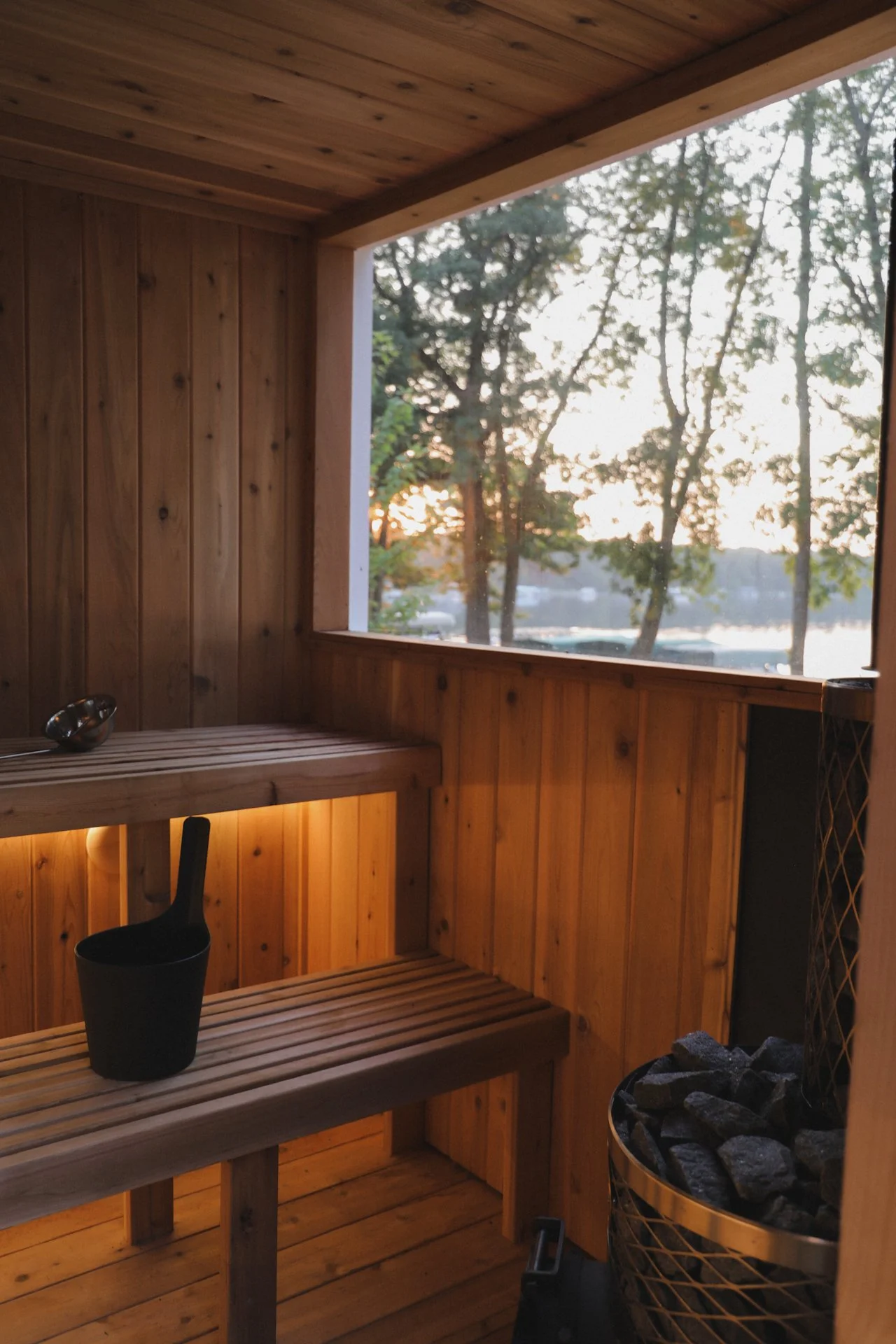 Interior of a wooden sauna with a bench, a black ladle, and a heater with rocks near a large window overlooking trees during sunset.