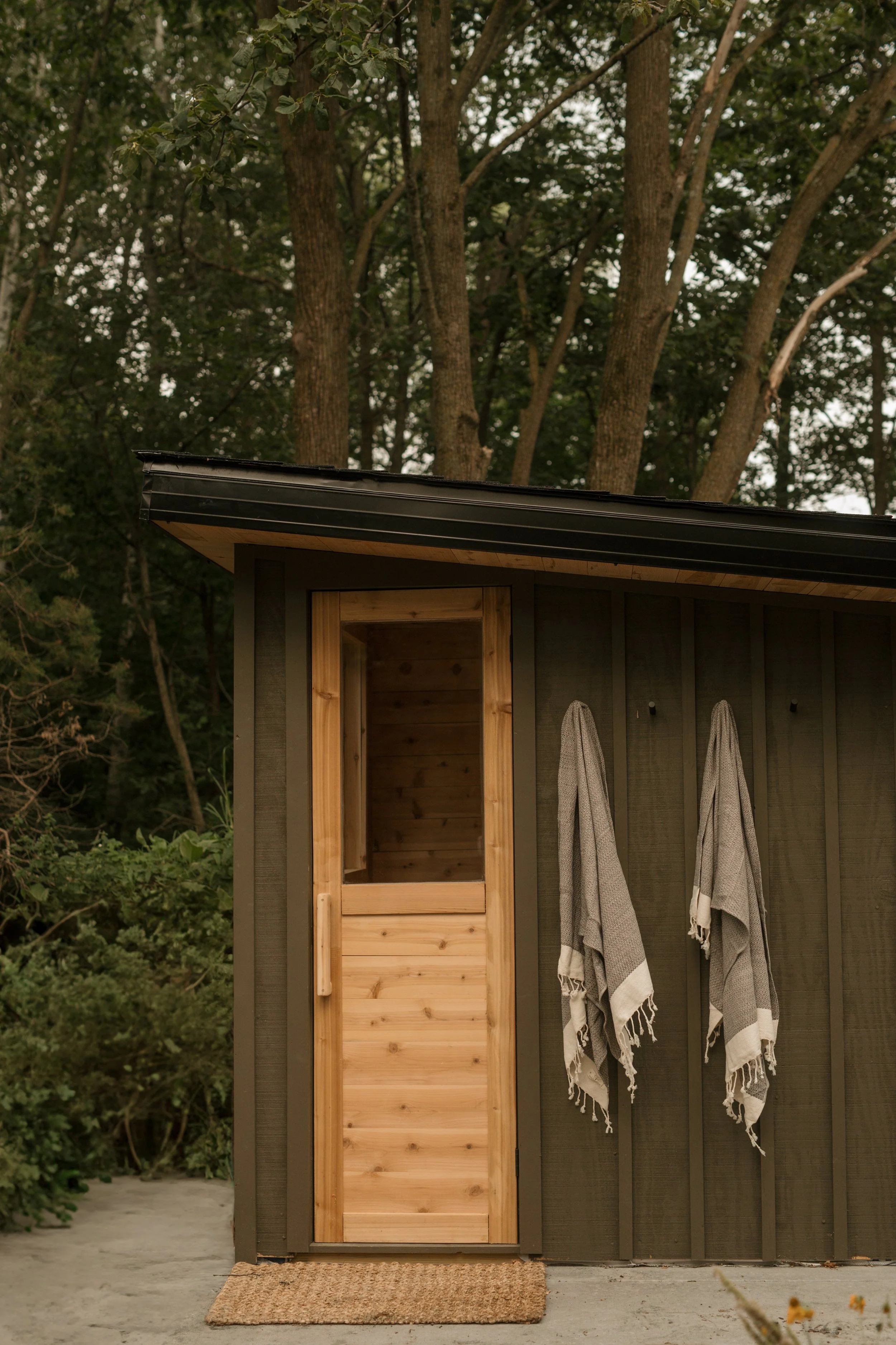 A small outdoor shower with a wooden door and two hanging towels, set against a dark wall, with trees in the background.