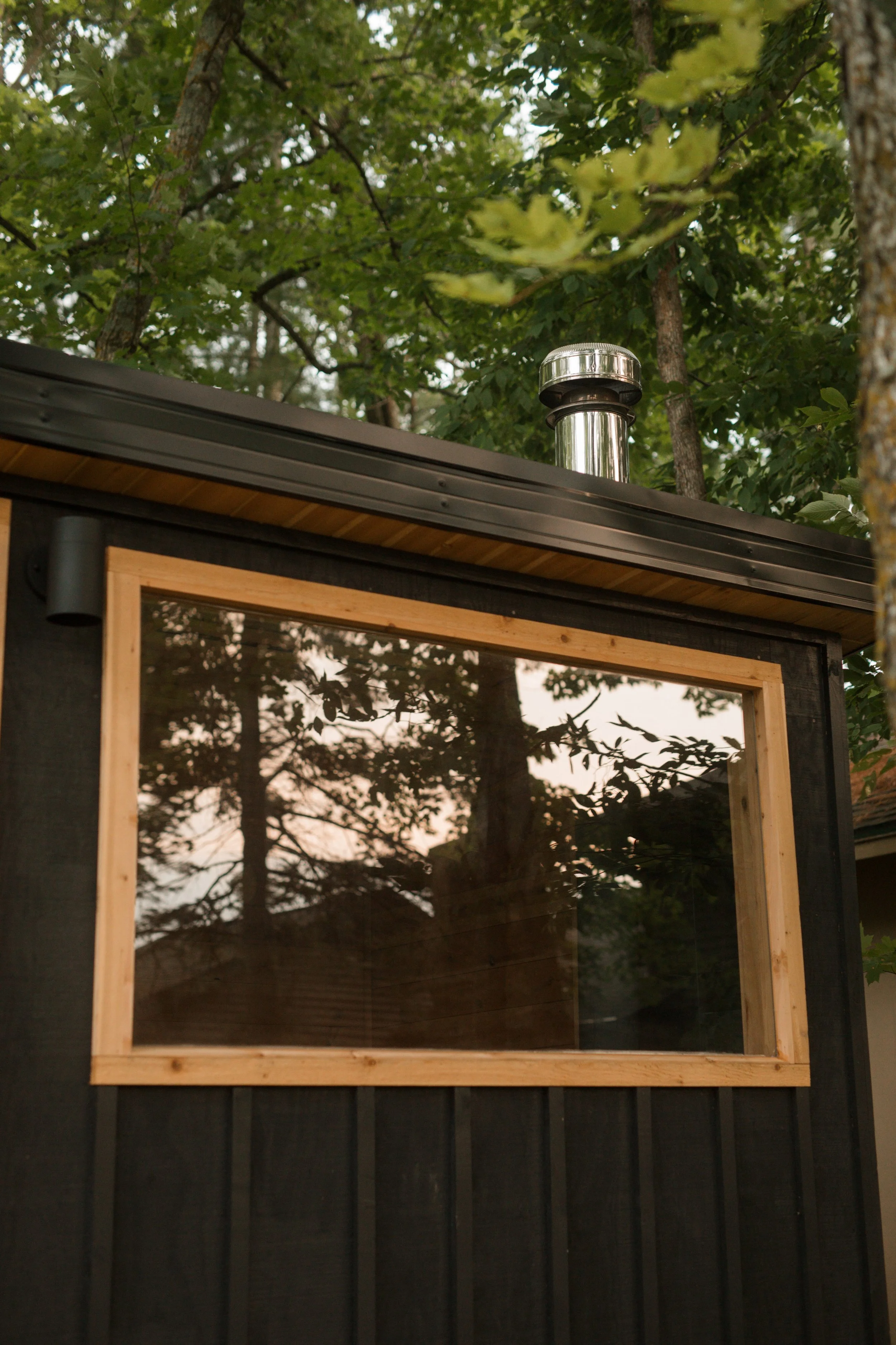 Close-up view of a black wooden structure with a window that has a wooden frame, set amidst trees with green leaves. Reflection of trees can be seen in the window, and a metal chimney pipe protrudes from the roof.