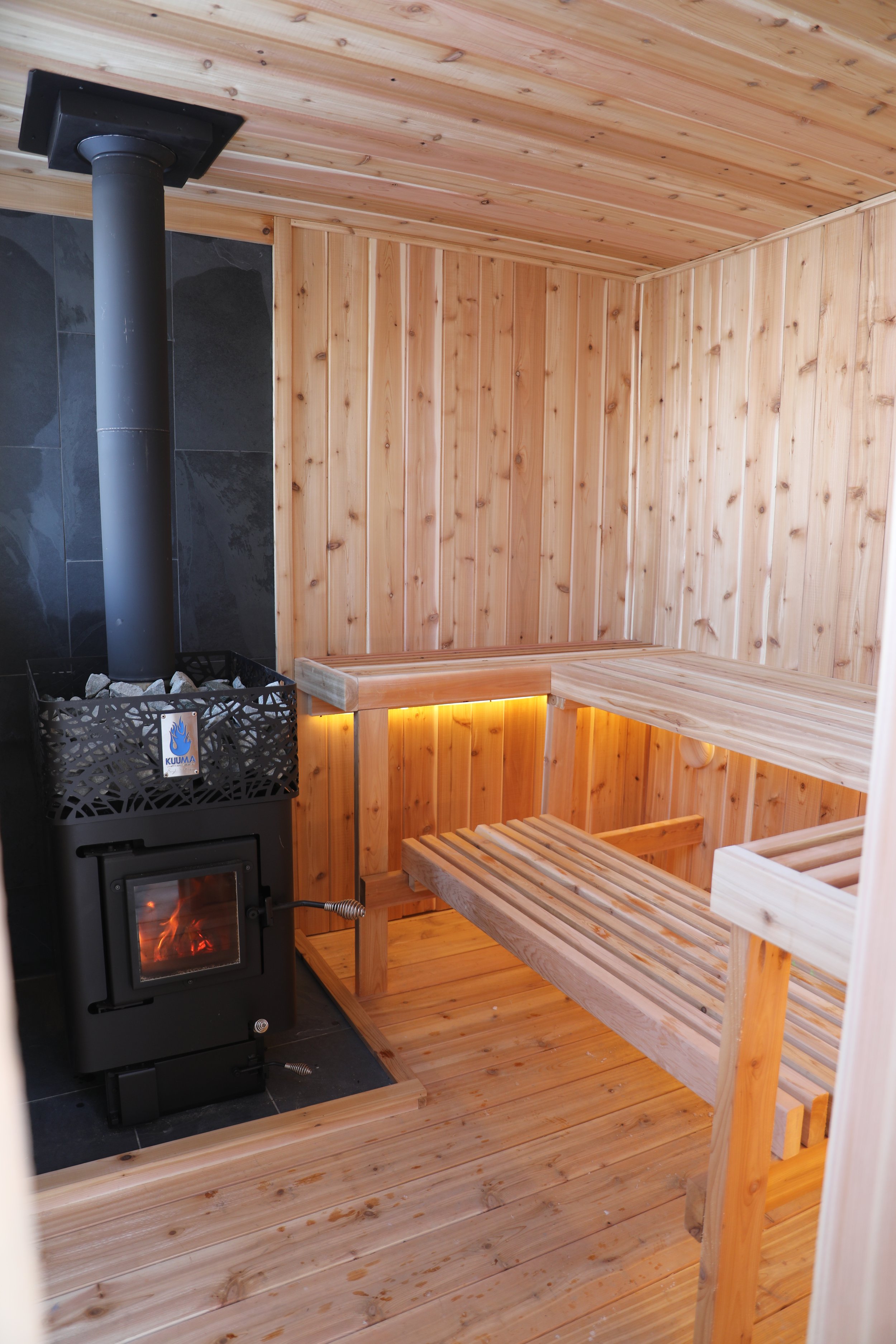 A wooden sauna room featuring a wood interior, a bench, and a black wood-burning stove with a glass door showing a fire inside.