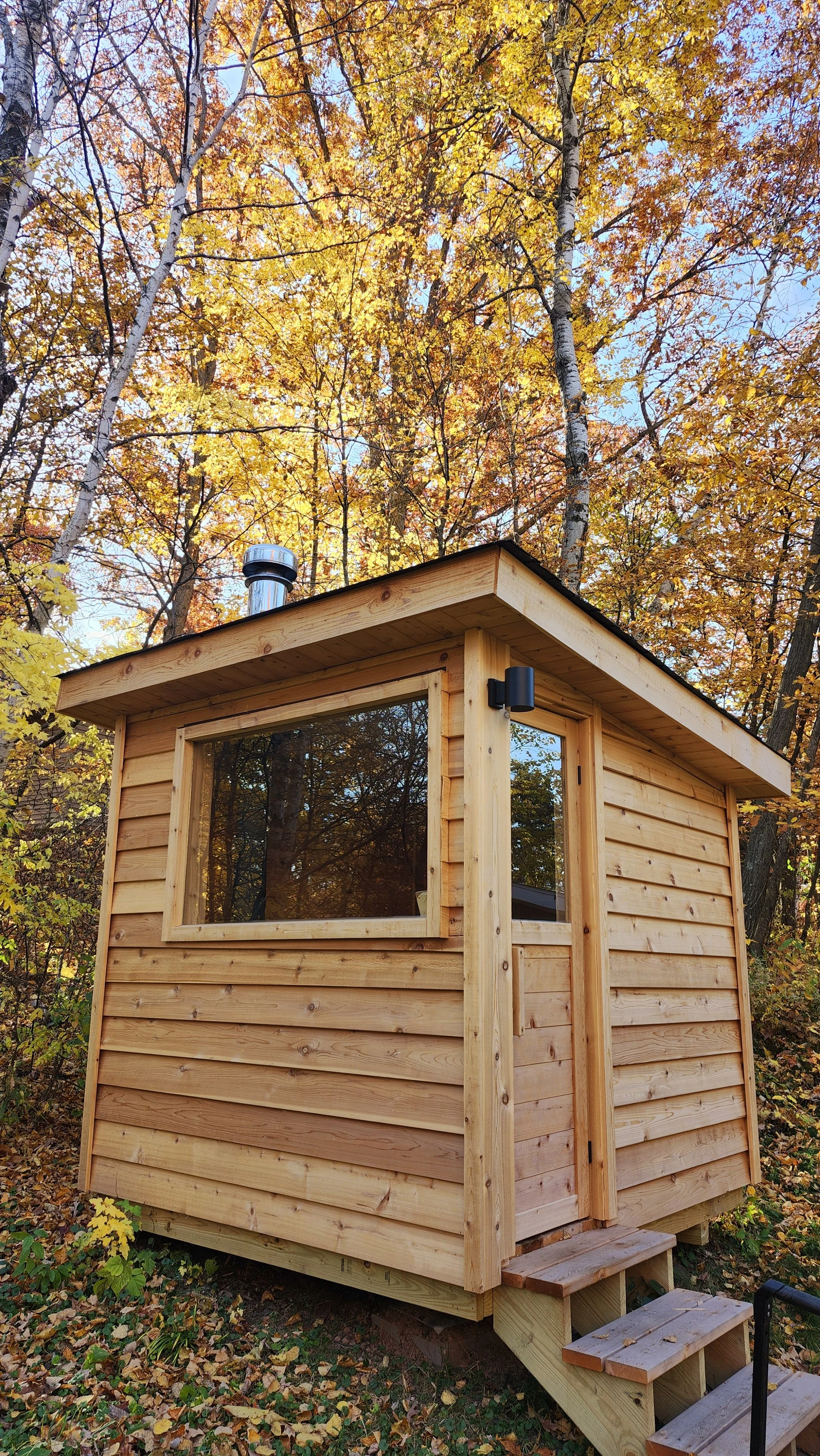 Small wooden cabin with natural wood siding, located in a forest with colorful autumn leaves and tall trees.
