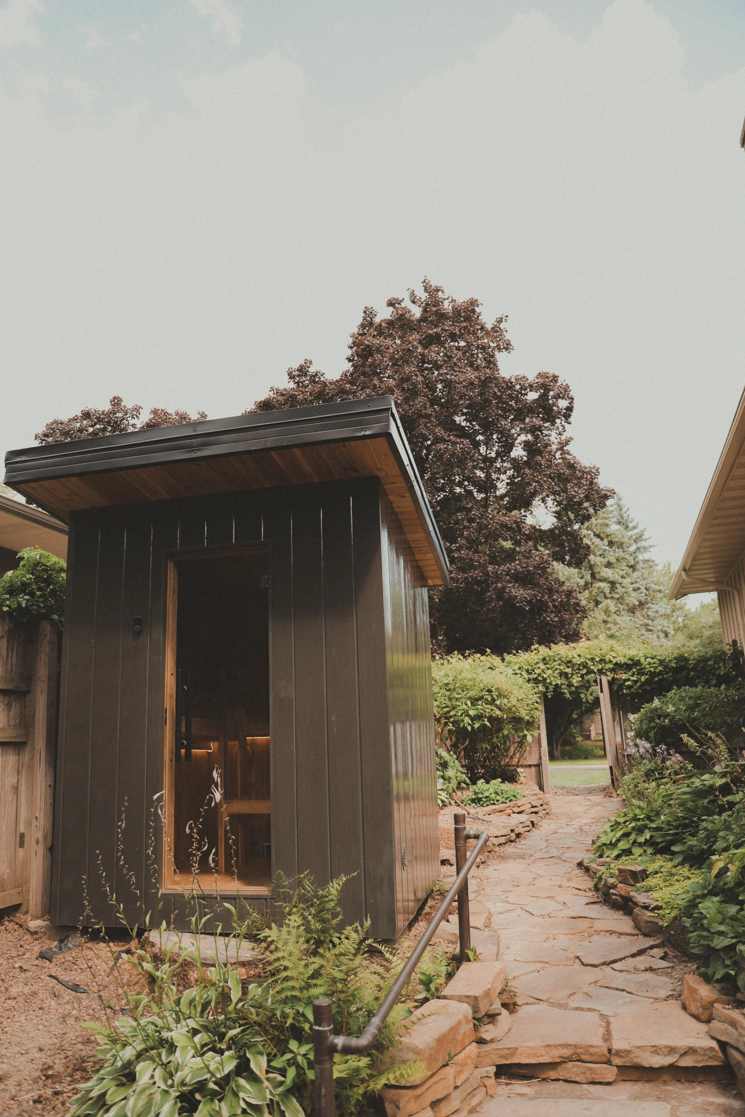 A small sauna in a garden with a stone pathway and lush green plants and trees in the background.