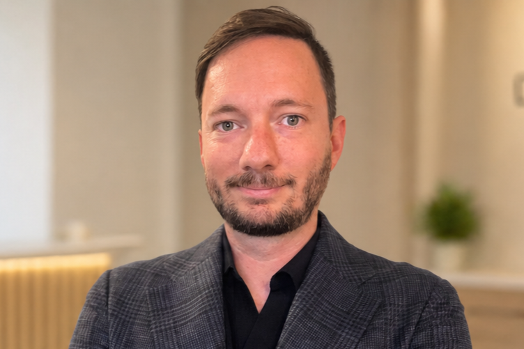 A man with short brown hair, beard, and blue eyes wearing a dark checked blazer and black shirt, standing indoors with a blurred background of a beige wall and a small green plant.